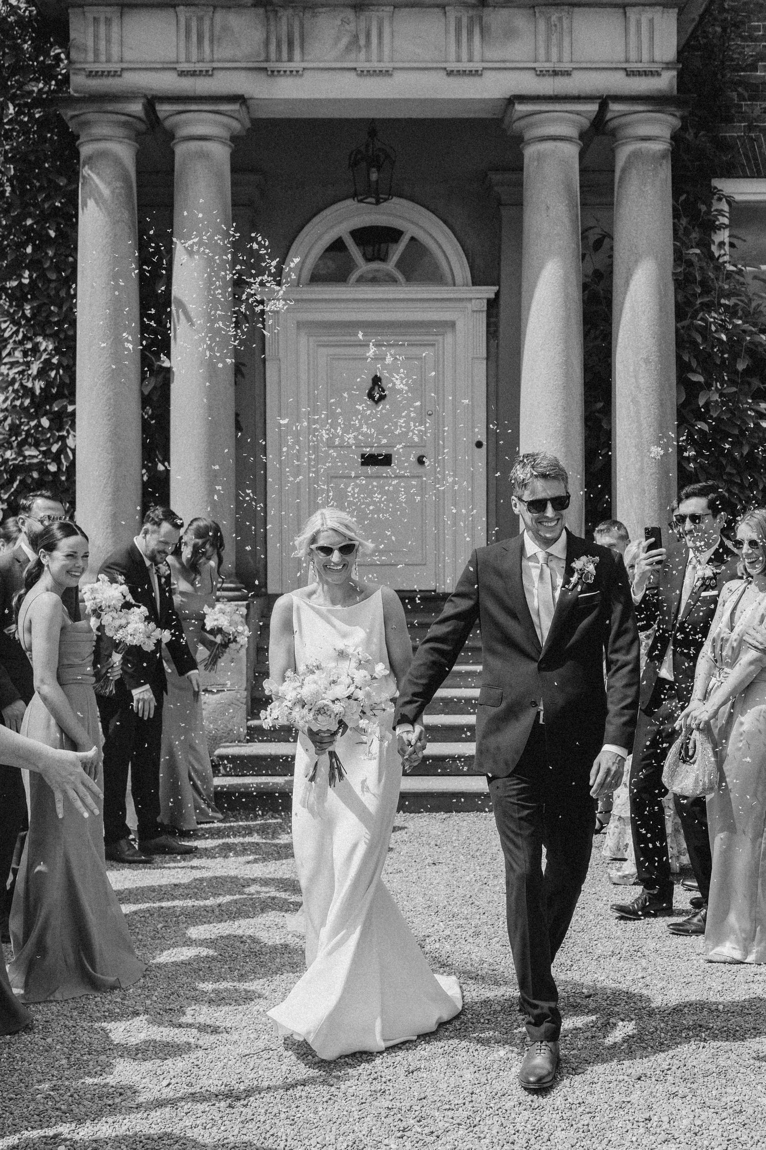Newlyweds walk through confetti shower past columned venue entrance surrounded by cheering wedding guests.