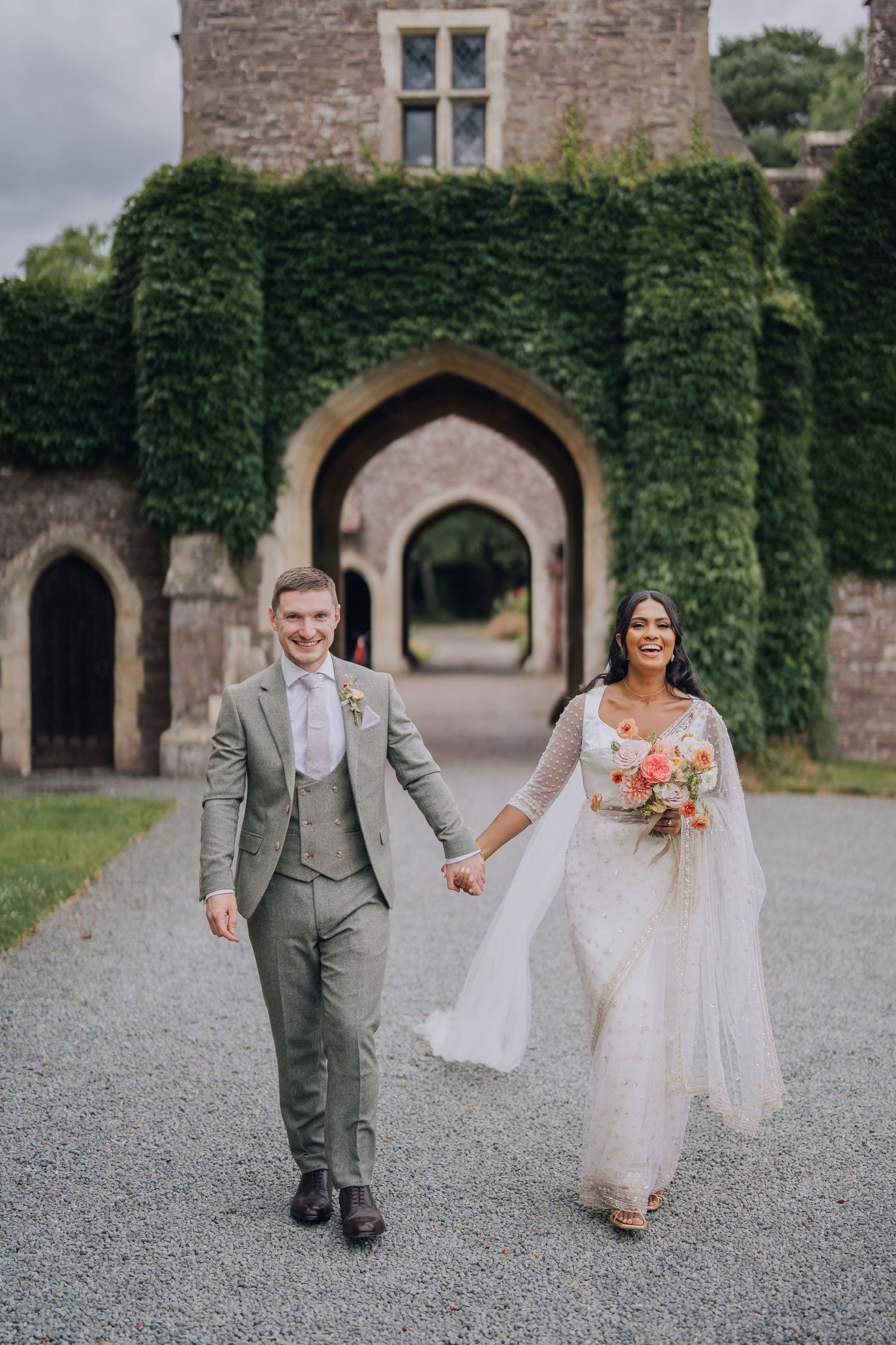 Joyful bride and groom walking hand in hand before ivy-covered castle archway, bride in lace gown with bouquet.