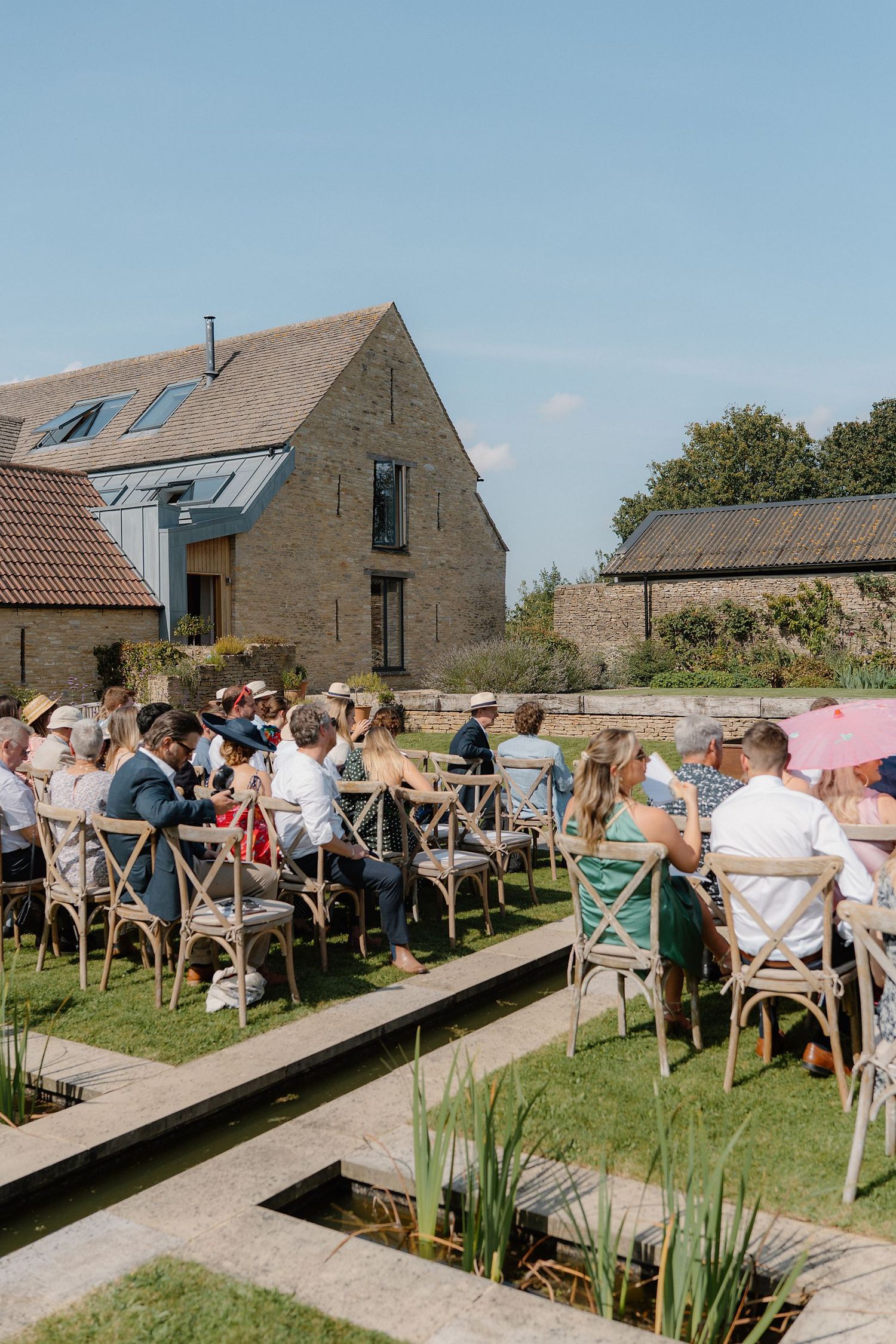 Wedding guests seated outdoors in sunshine at rustic stone barn venue ceremony.