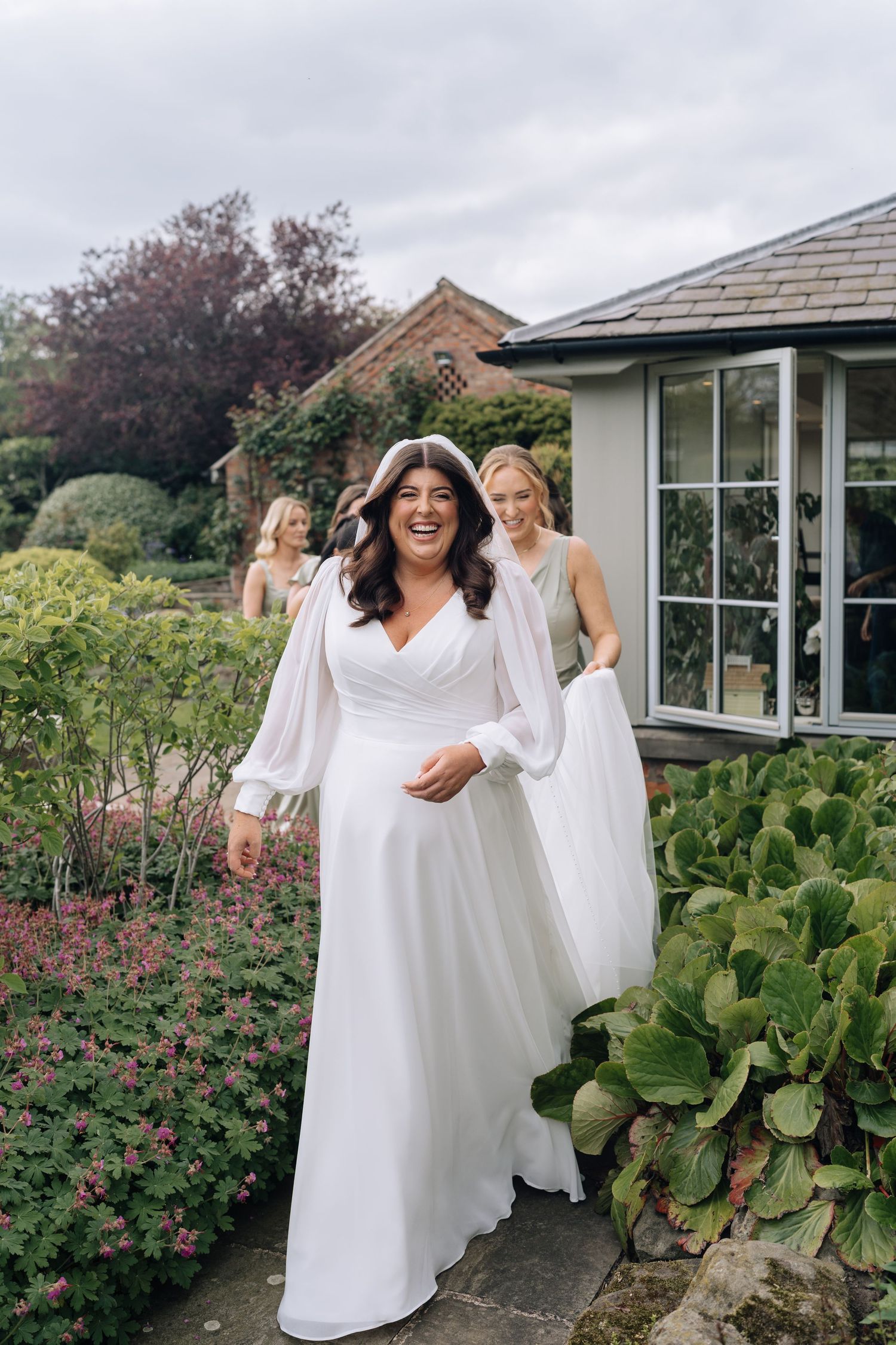 Bride in white long-sleeve gown laughs in garden, bridesmaids in sage green dresses assist with train.