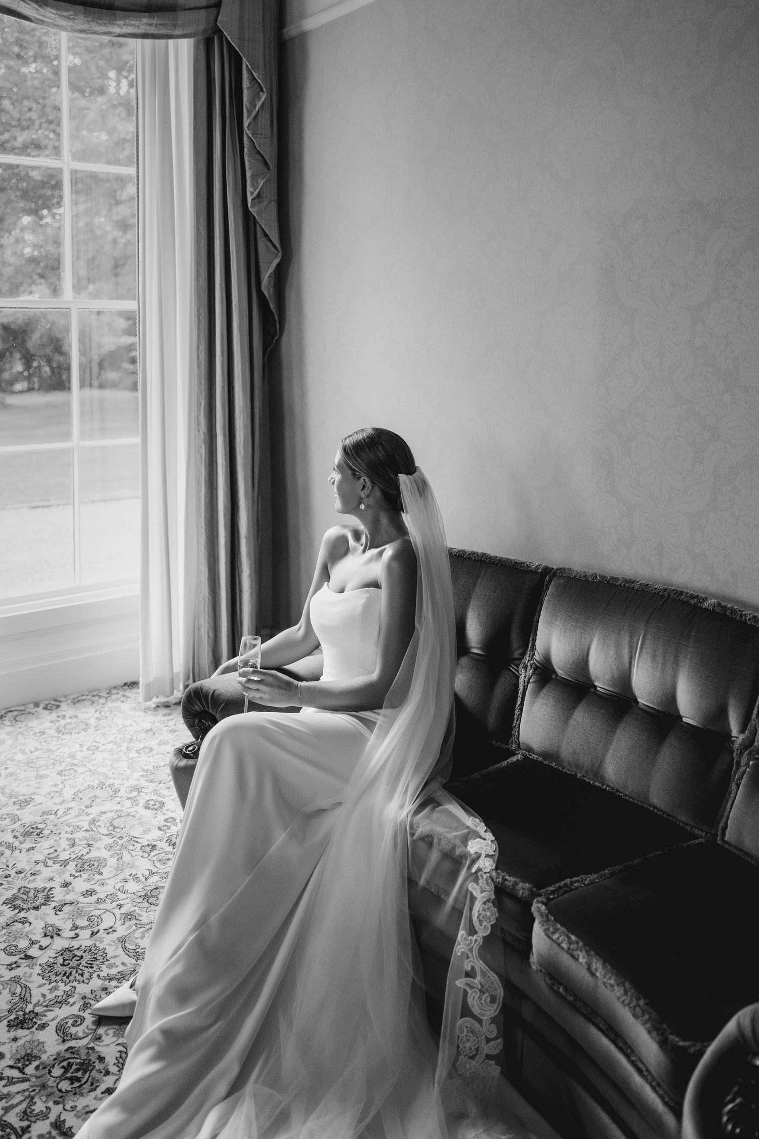 Black and white photo of a bride in a white gown and veil sitting on a leather sofa gazing out a large window.