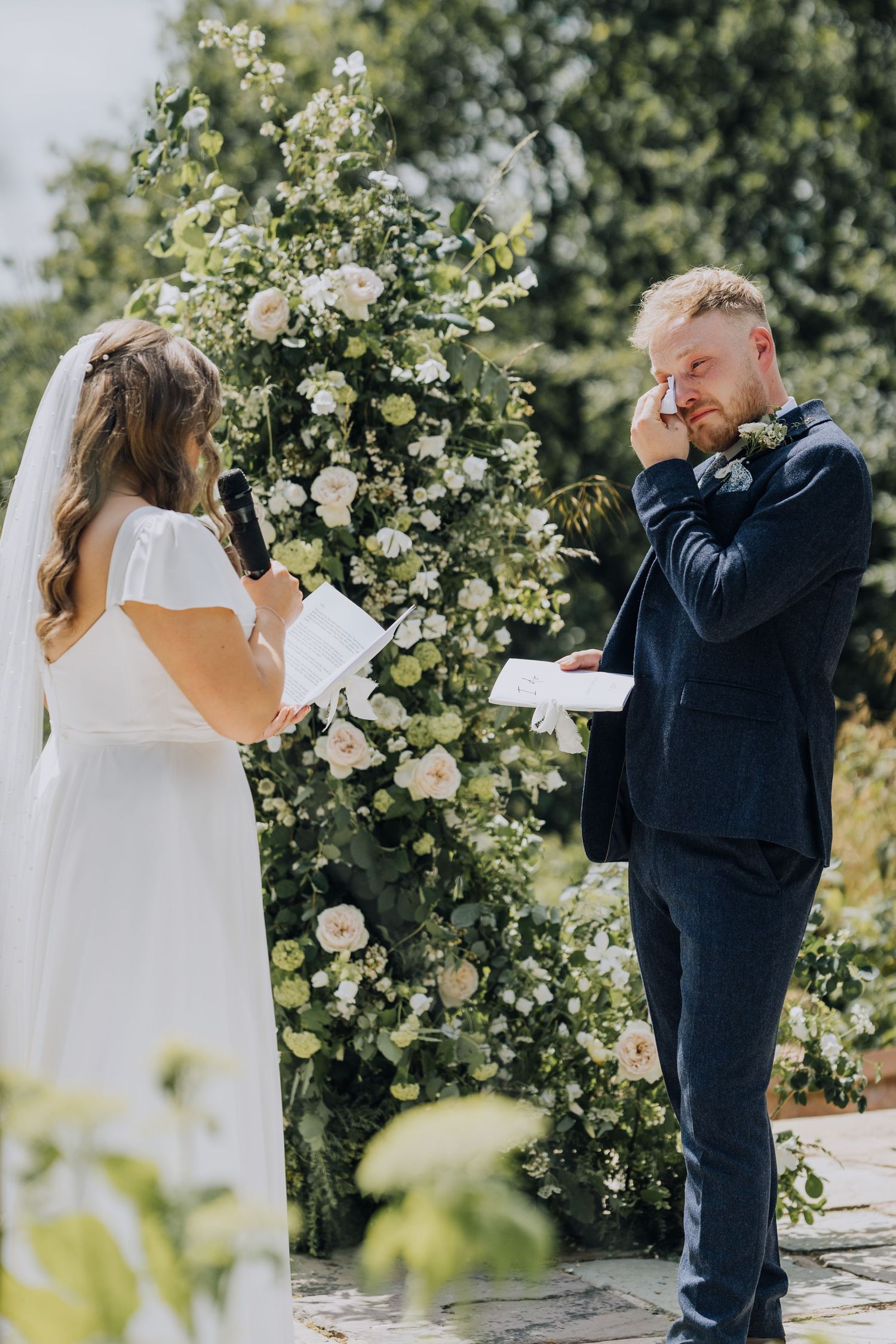 Tearful groom wipes eyes as bride reads vows at floral arch outdoor wedding ceremony.