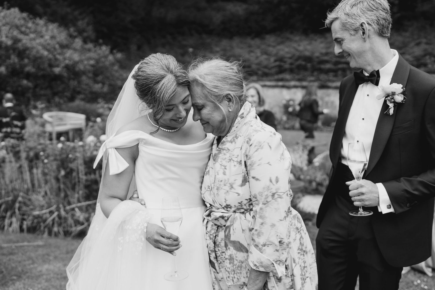 Bride sharing tender moment with elderly grandmother and groom in black and white wedding photo.