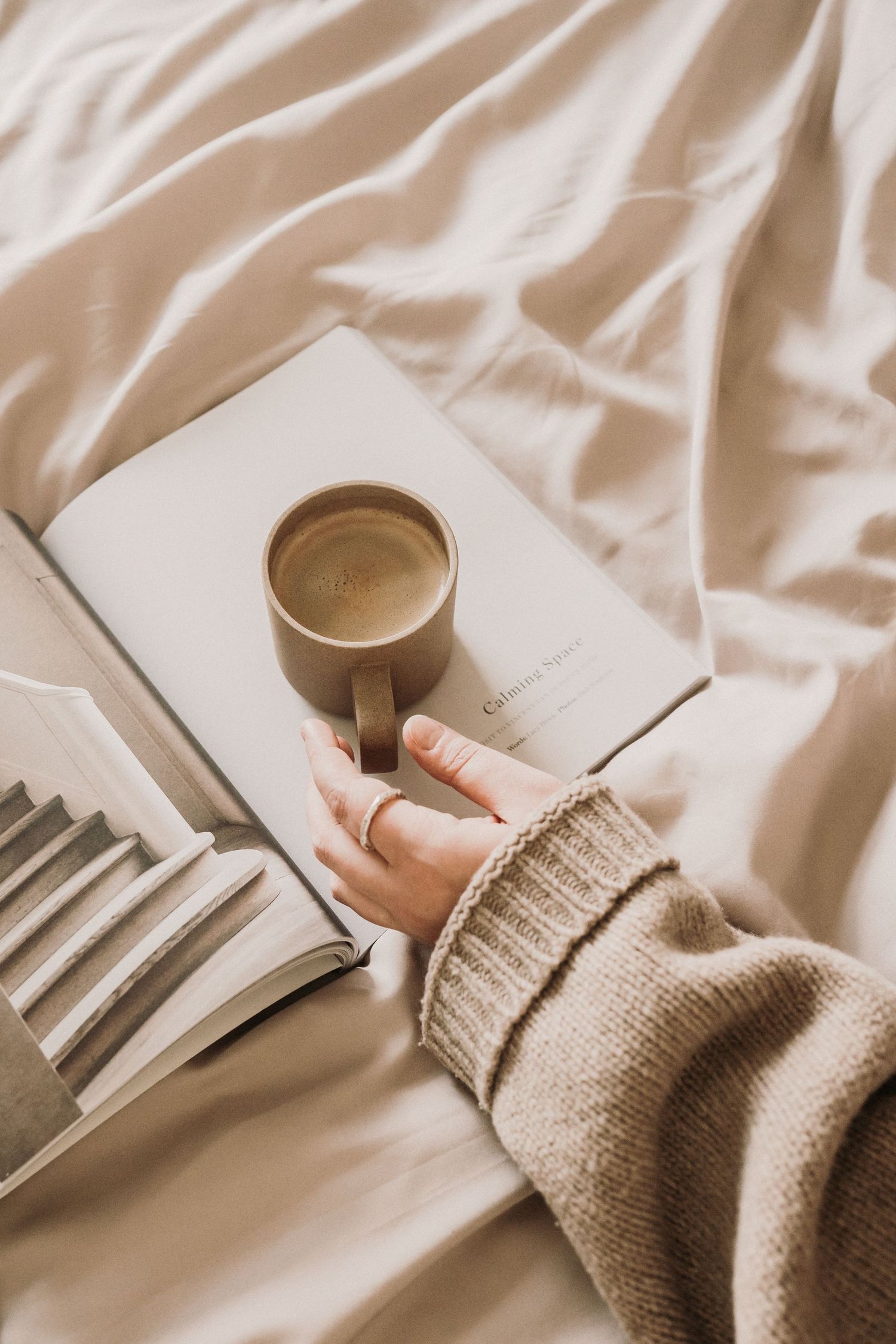 A person in a beige sweater holding a coffee cup over an open book on white bed linen.