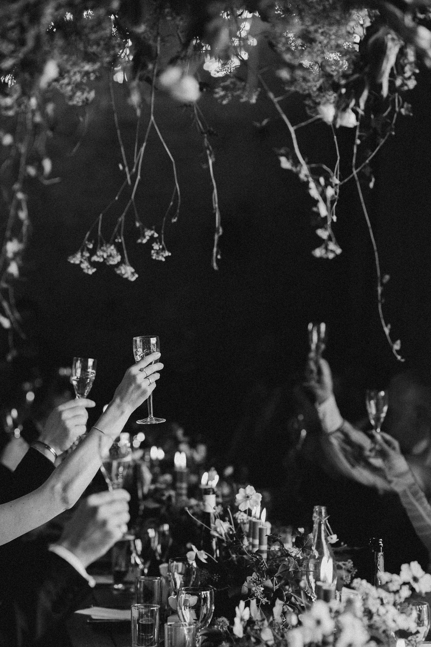 Black and white photo of wedding guests raising champagne glasses in toast beneath hanging floral ceiling installation.