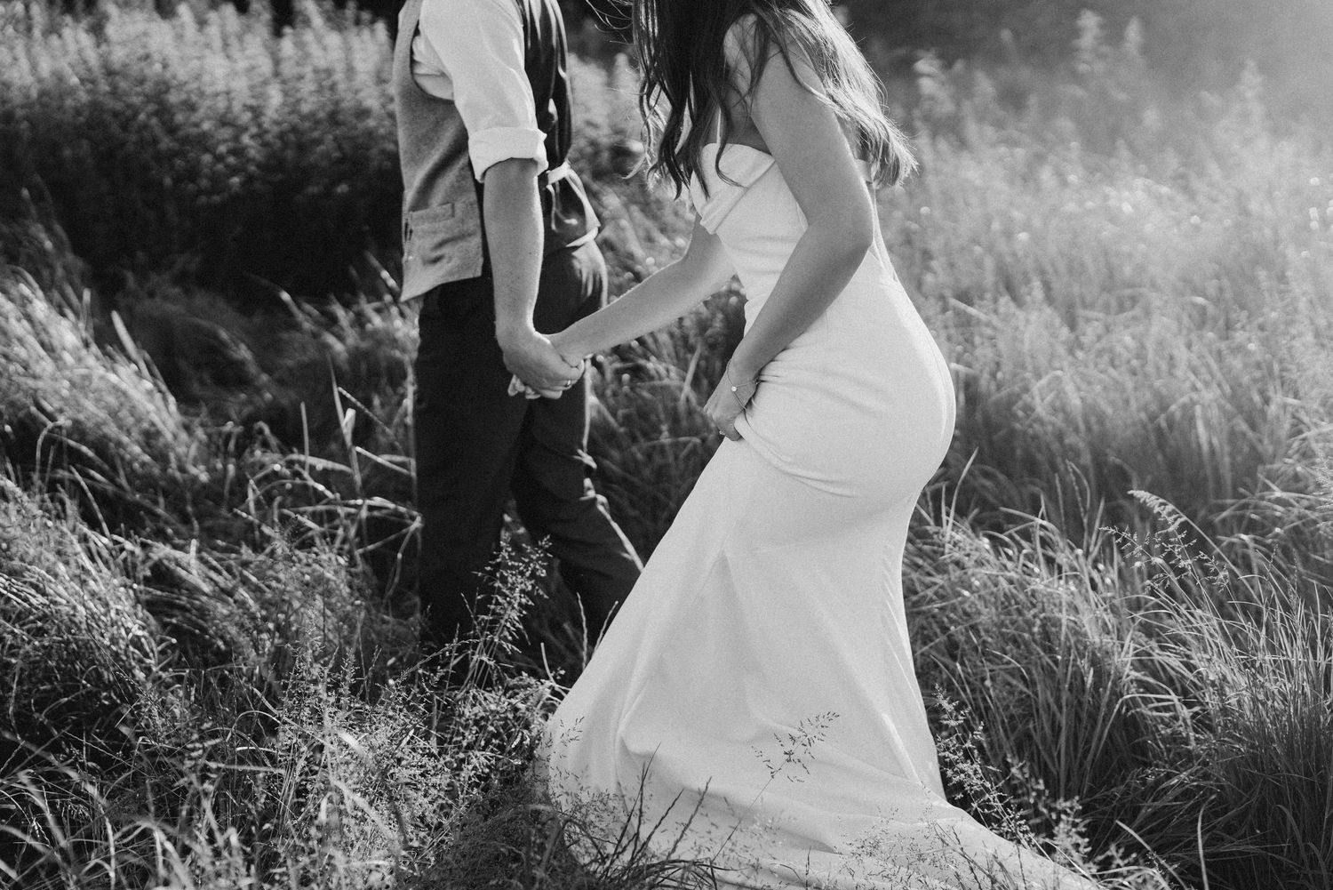 Black and white romantic photo of bride in elegant white dress walking hand in hand with groom in field.
