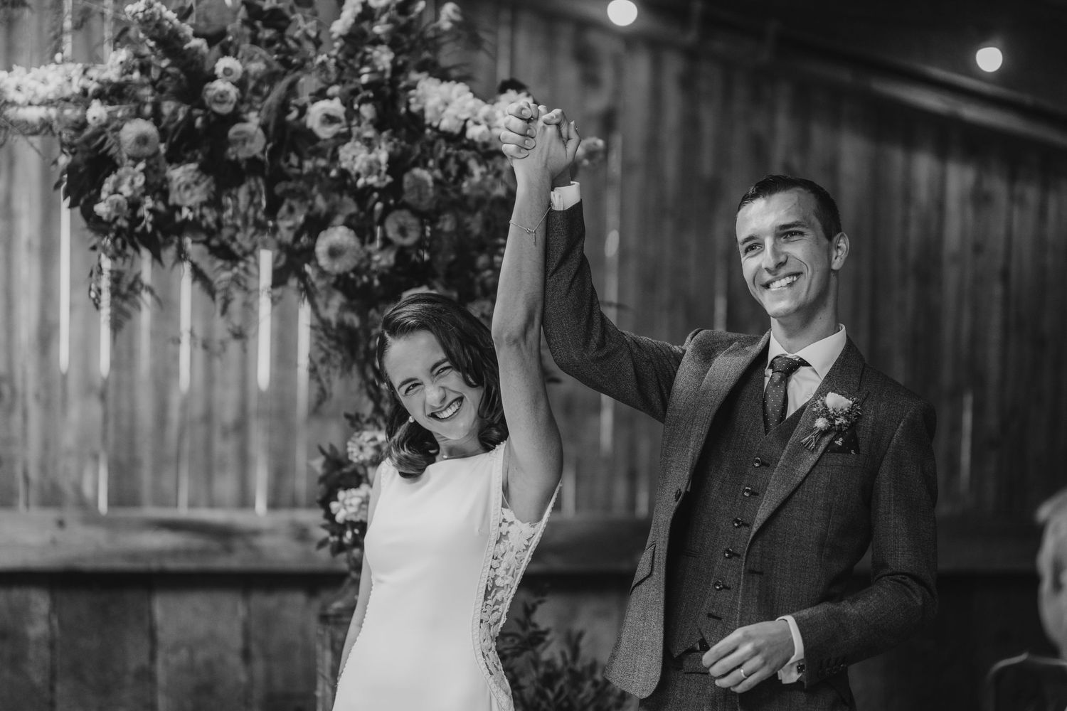Black and white photo of newlyweds celebrating with raised hands before floral arch in barn.
