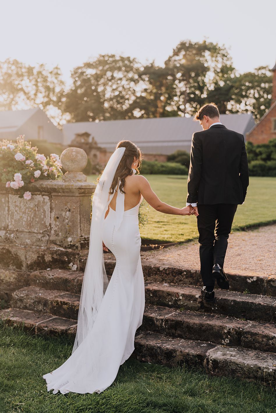Bride in flowing white gown and veil holds grooms hand walking up stone steps in golden sunset light.