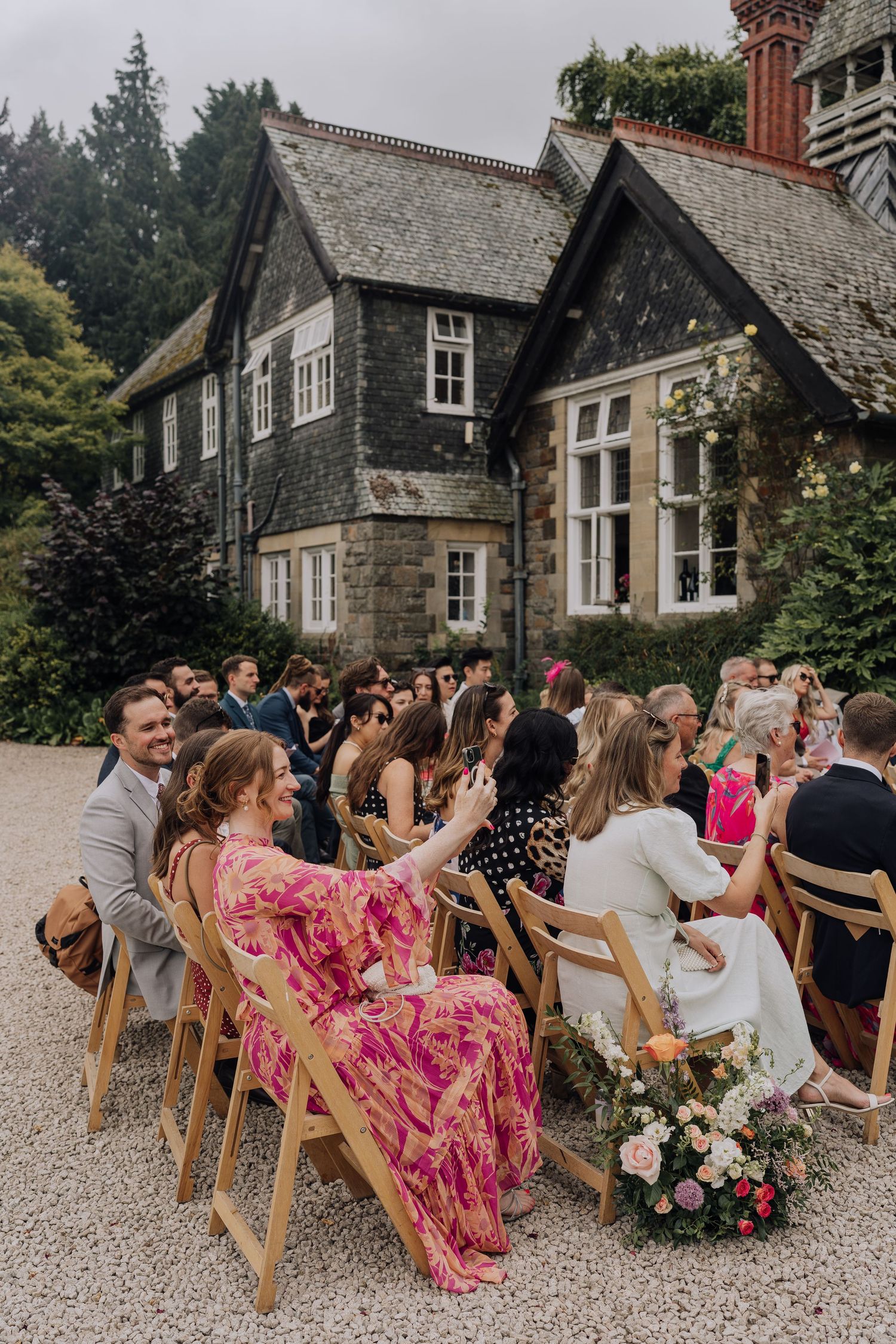 Wedding guests seated in wooden chairs outdoors for ceremony in front of charming stone manor house.