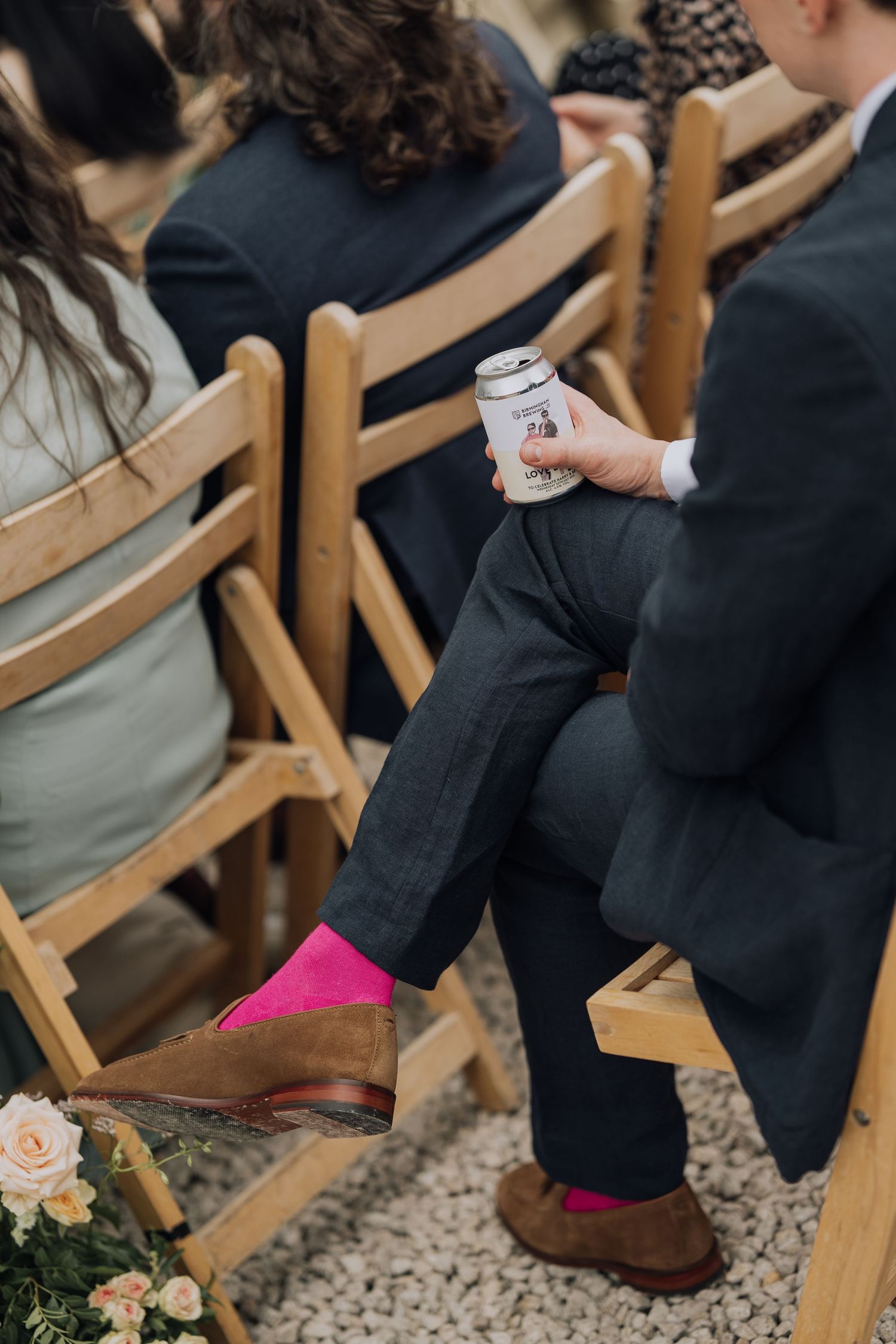 Wedding guest with bright pink socks and brown loafers holding beverage can during outdoor ceremony.