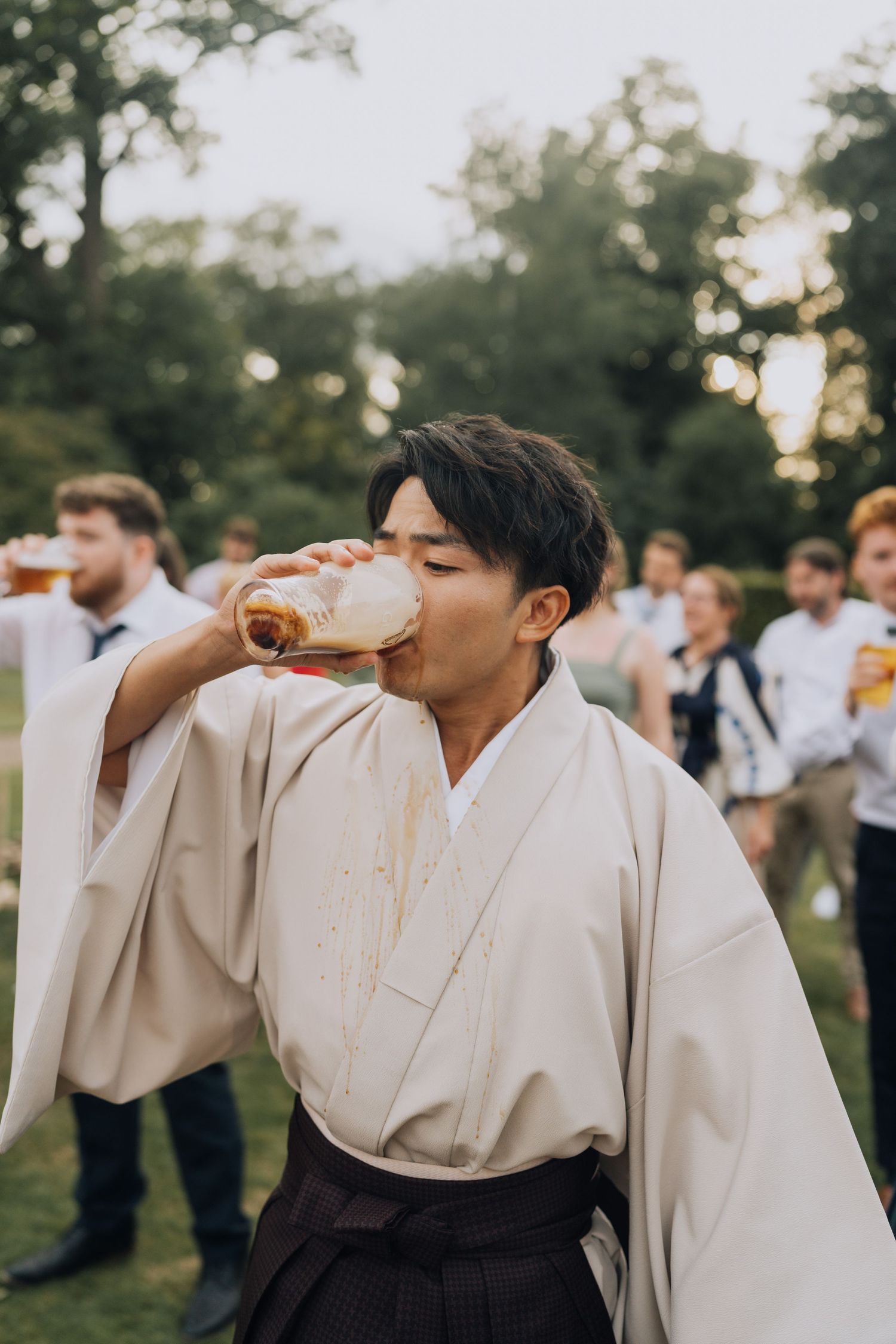 Man in traditional Japanese kimono drinking beer at outdoor garden party with guests in background.