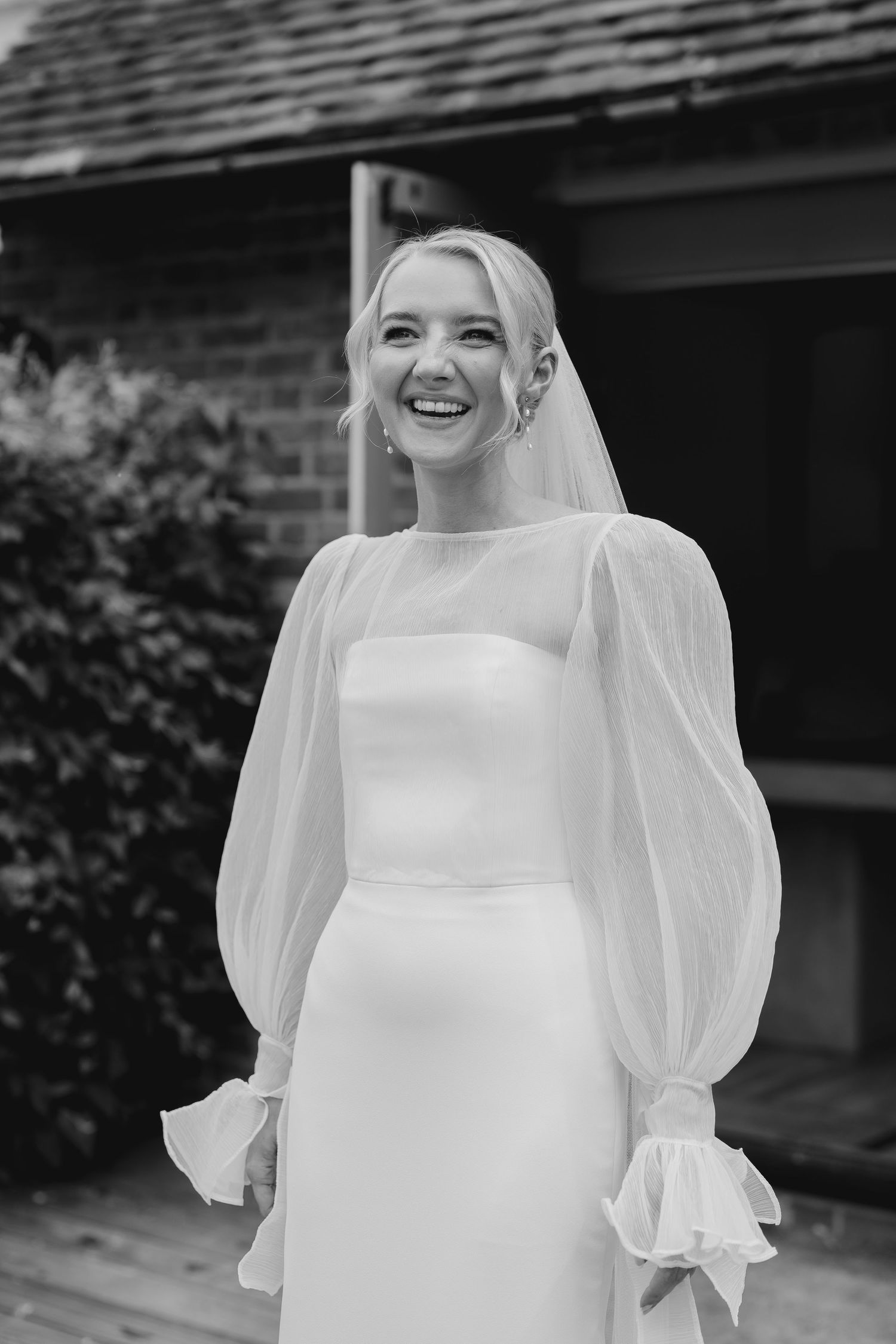 Black and white photo of a laughing bride in a white gown with sheer puff sleeves and veil outdoors.