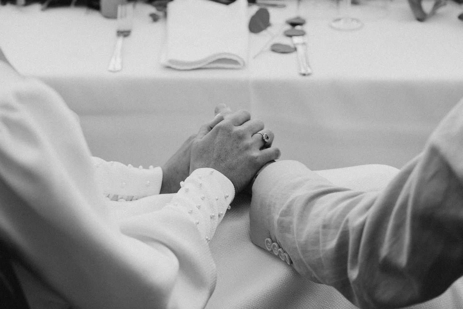 Black and white close-up of newlyweds holding hands tenderly at the wedding reception table.