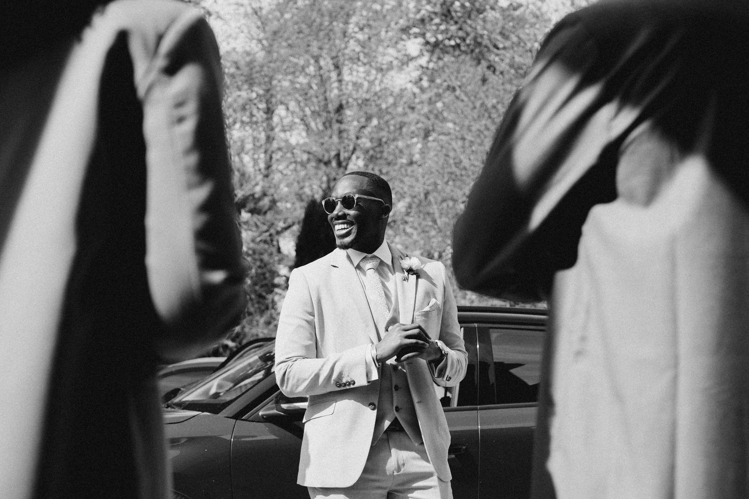 Smiling man in light suit and sunglasses standing by a car in a black and white outdoor photo.
