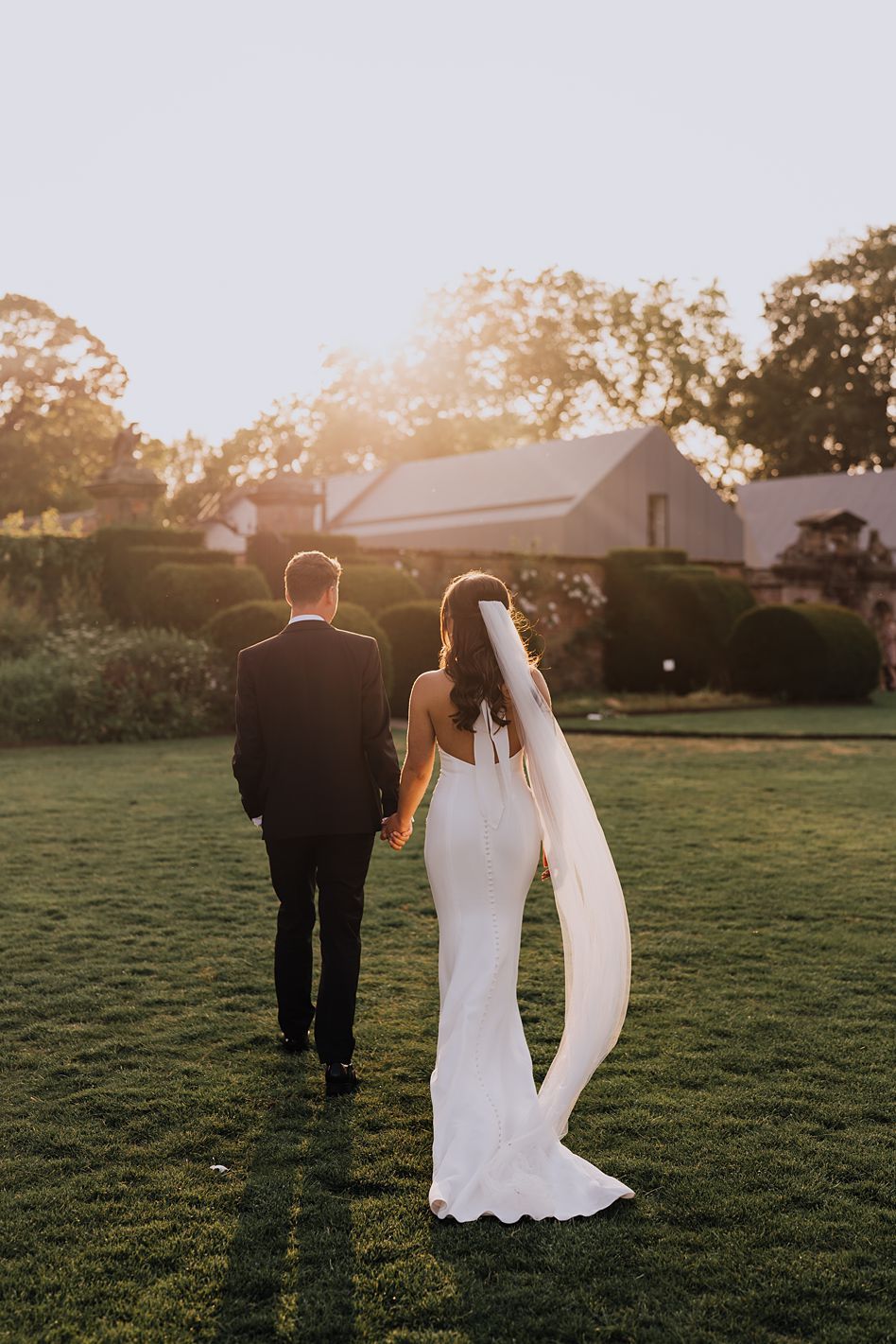 Bride in backless gown with long veil and groom walking hand in hand toward a sunlit estate.