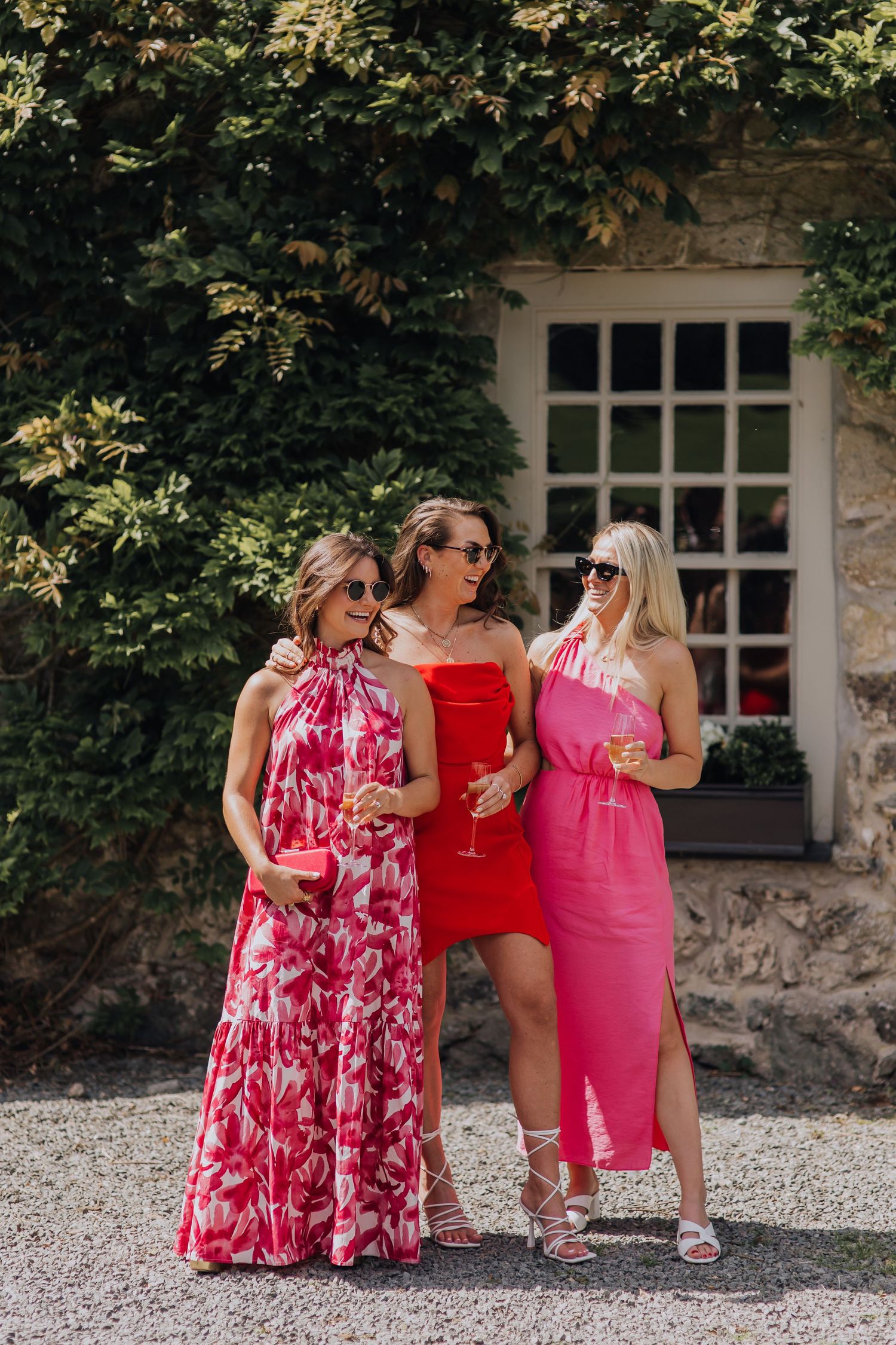 Three women in pink and red dresses holding champagne glasses outside a stone building at a wedding.