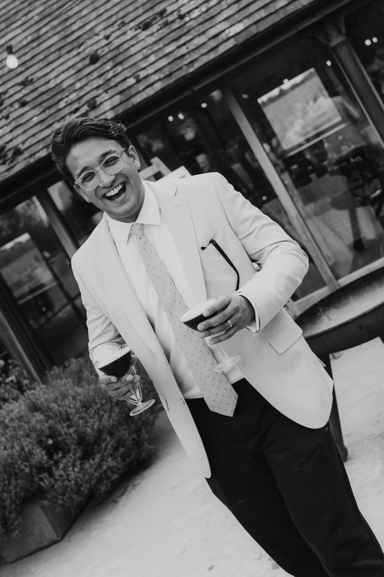 Smiling man in white blazer and tie holding a wine glass outdoors at a wedding celebration.