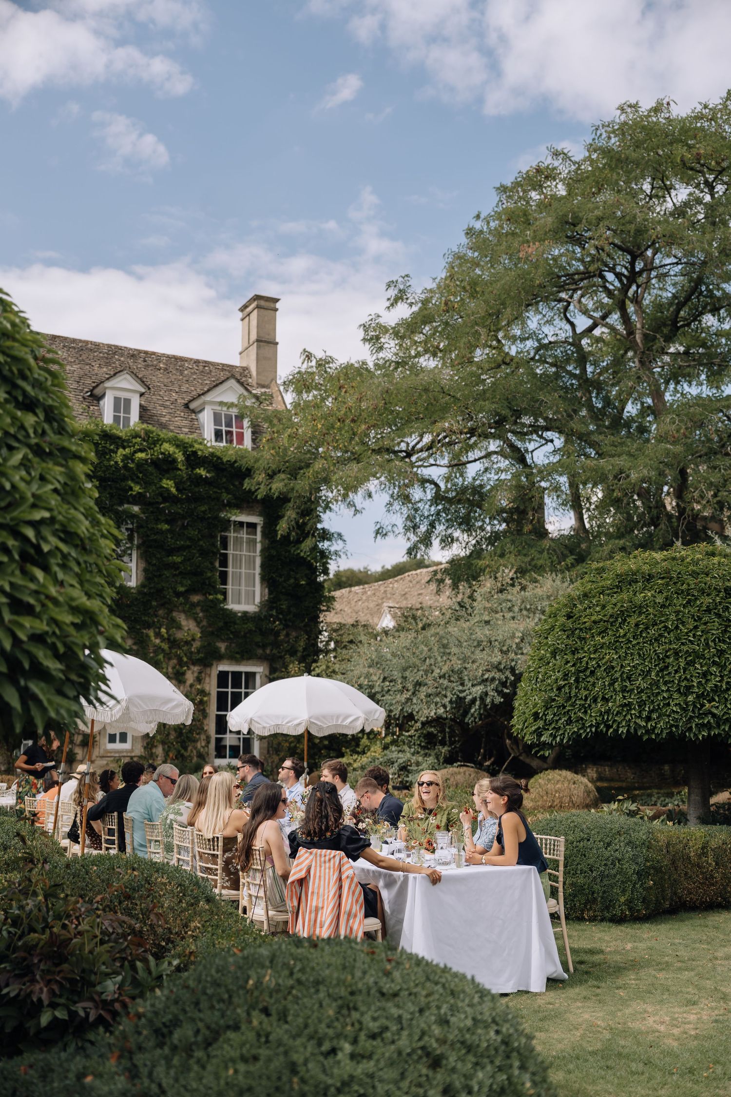 Guests gather for an elegant outdoor garden party at a ivy-covered stone manor house on a sunny day.