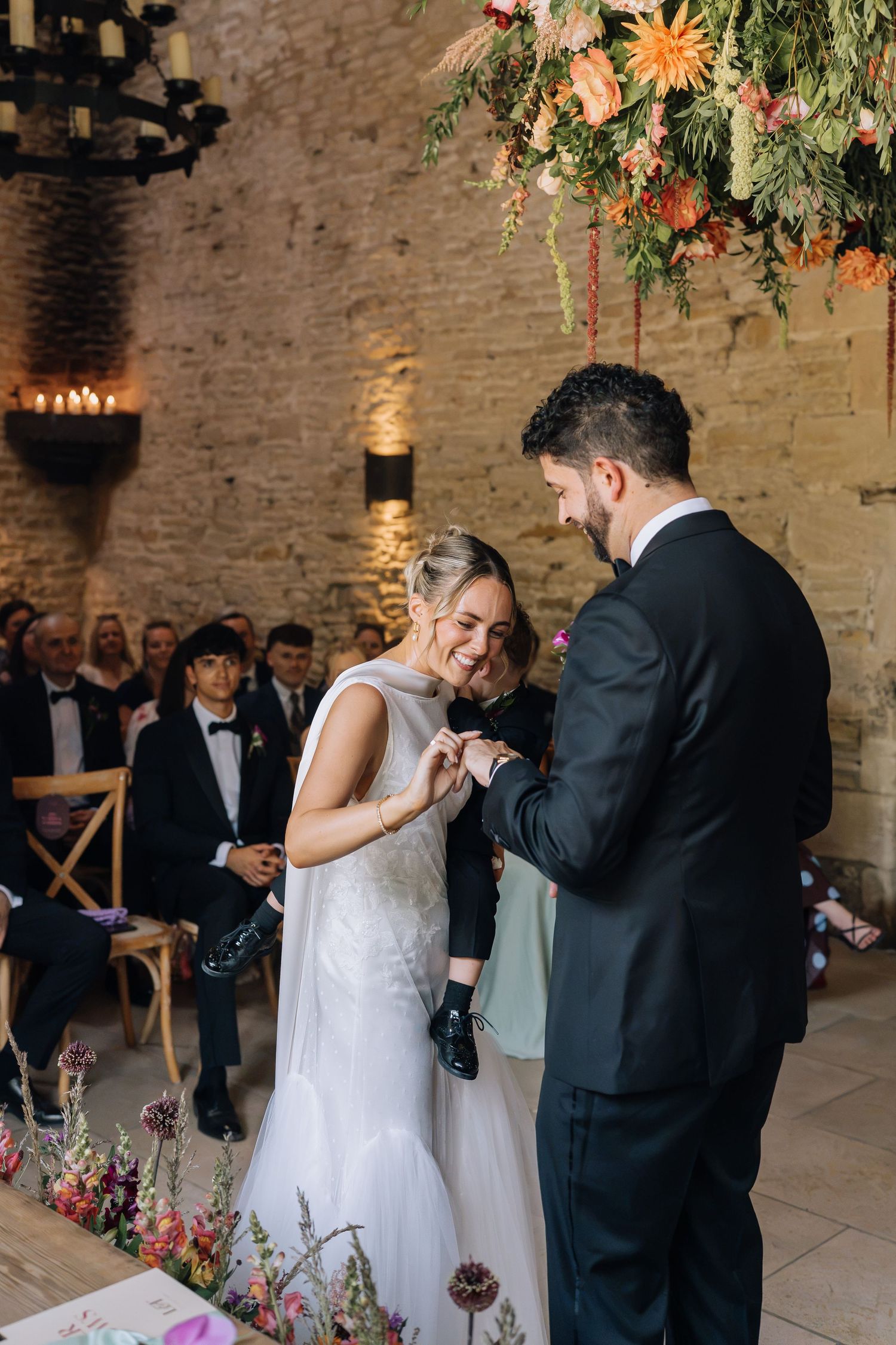 Bride and groom exchange rings during wedding ceremony in rustic stone venue with floral arrangements.