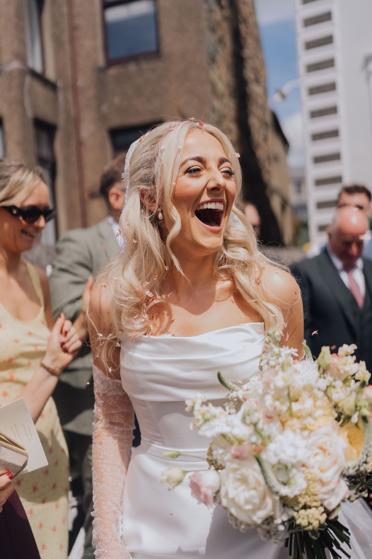 Joyful bride in white strapless gown laughing while holding a pastel bouquet during outdoor wedding confetti celebration.
