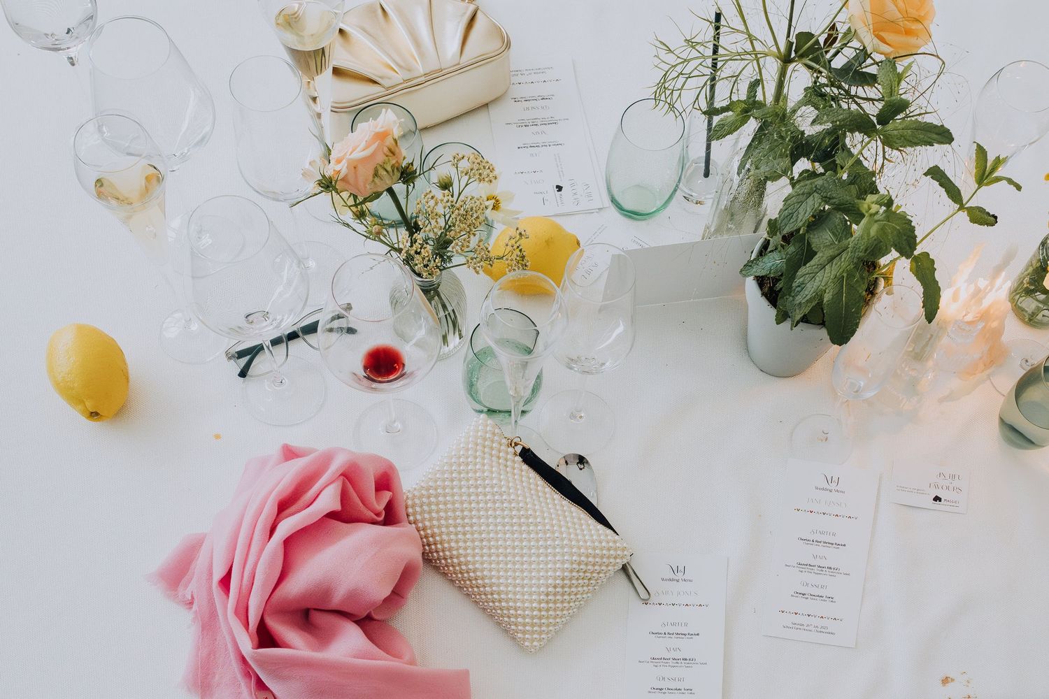 Elegant wedding table flatlay with yellow roses, lemons, pink ribbon, glassware, and floral arrangements on white linen.