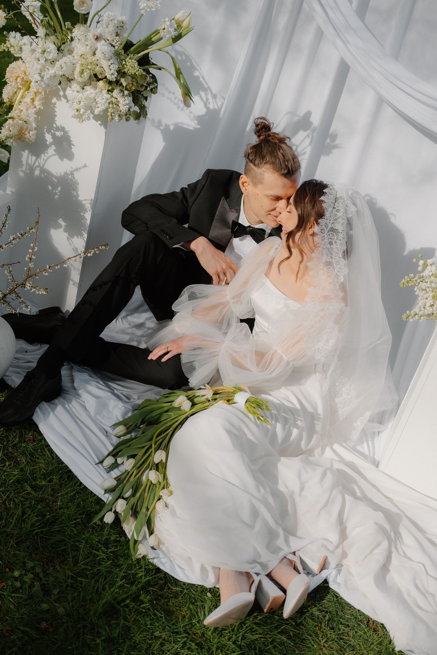Bride and groom sit together on grass surrounded by white tulips and flowing white draping.