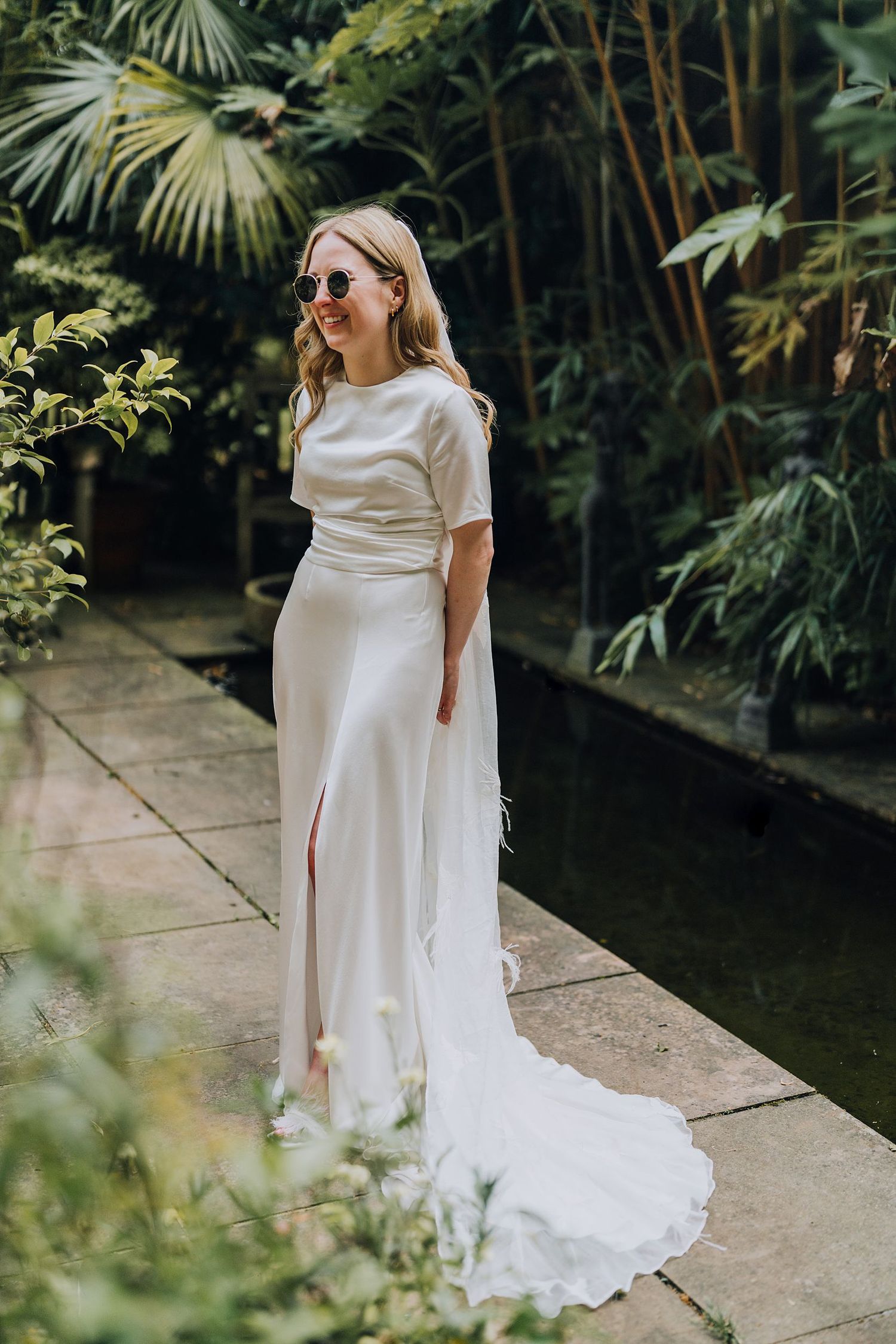 Stylish bride in white satin gown and sunglasses posing in lush tropical garden setting.
