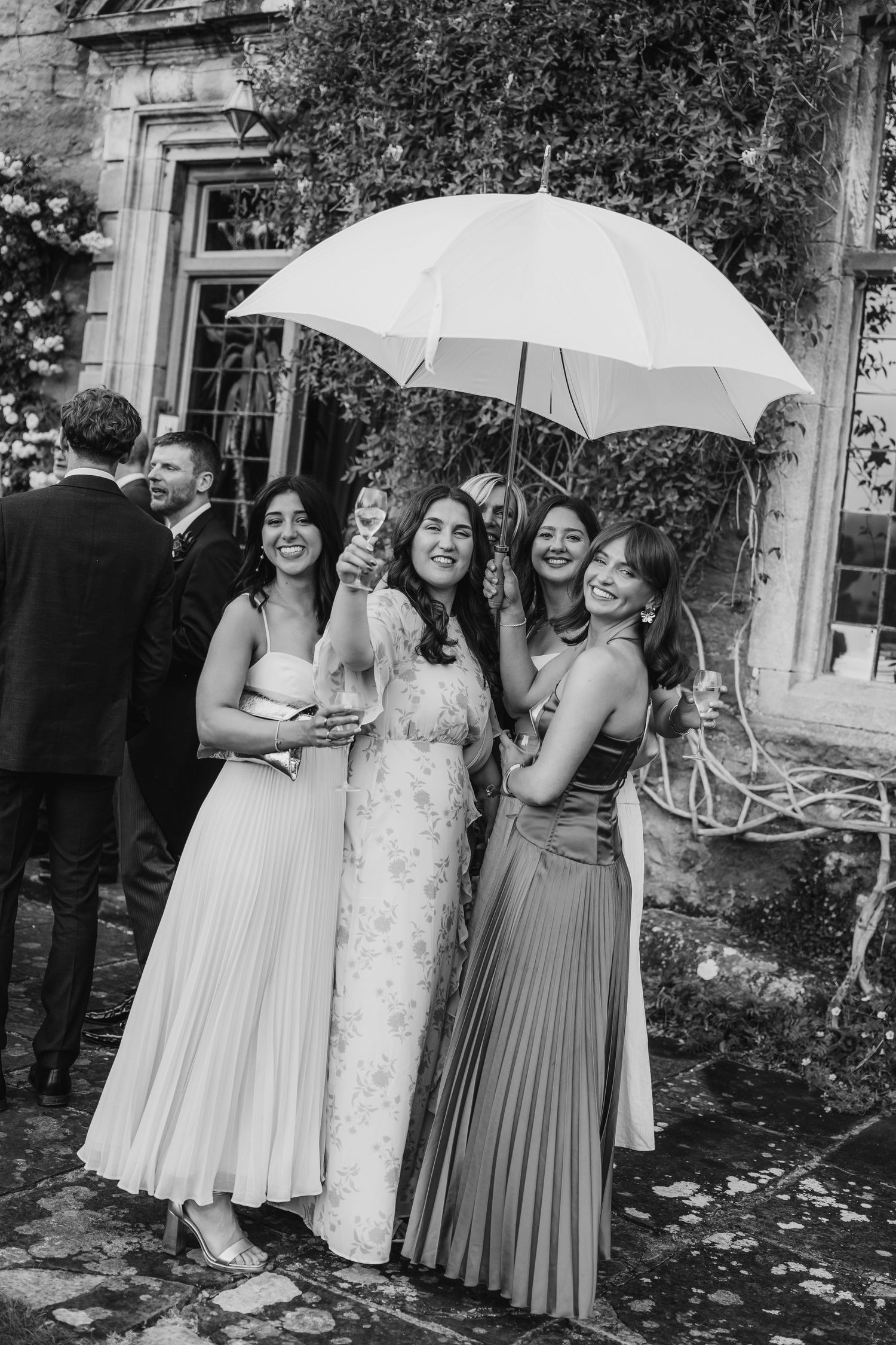 Black and white photo of four women sharing an umbrella and laughing at a rainy outdoor wedding.