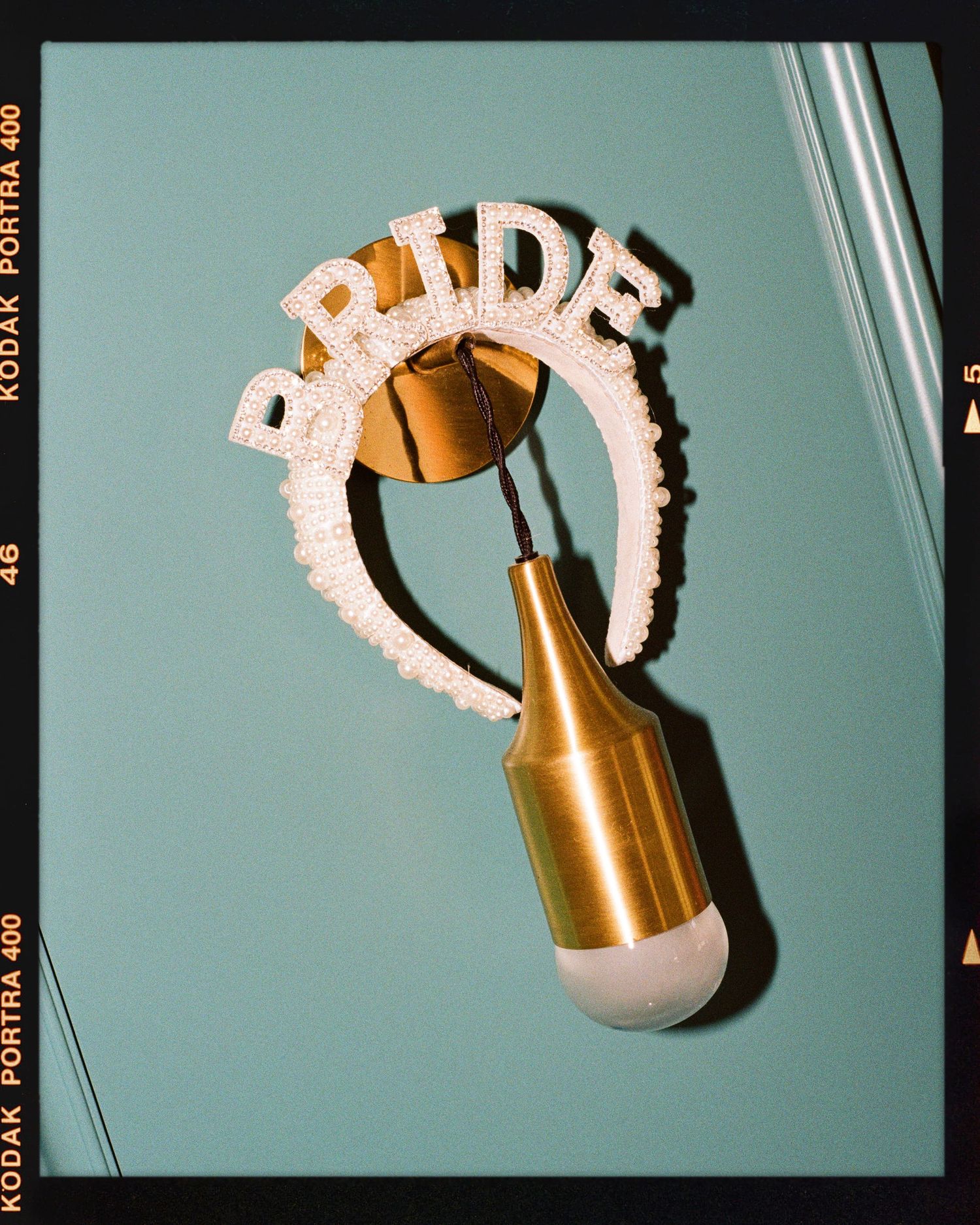 A pearl BRIDE headband displayed on a gold pendant light fixture against a teal wall, shot on Kodak Portra 400 film.