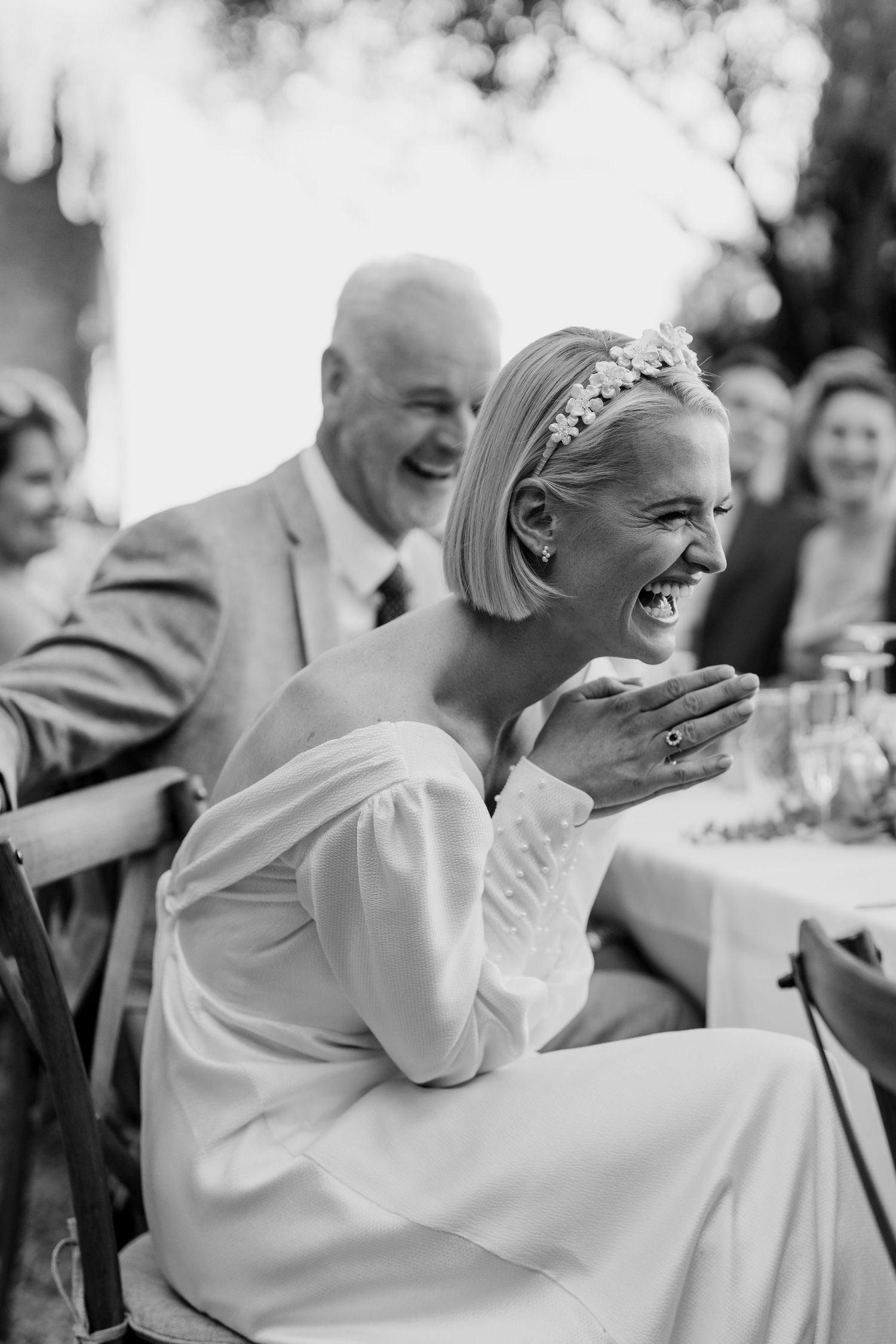 Laughing bride wearing floral headband at outdoor wedding reception in black and white photo.