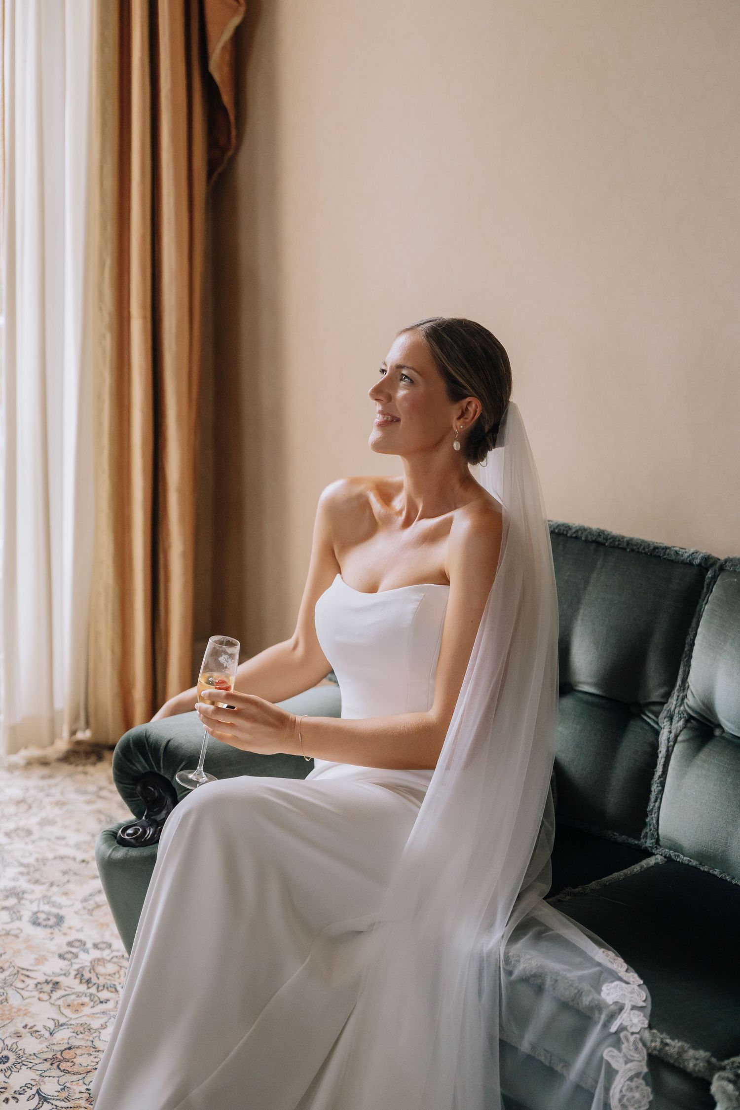 Elegant bride in white strapless gown and veil sits on green sofa holding champagne glass, smiling near window.