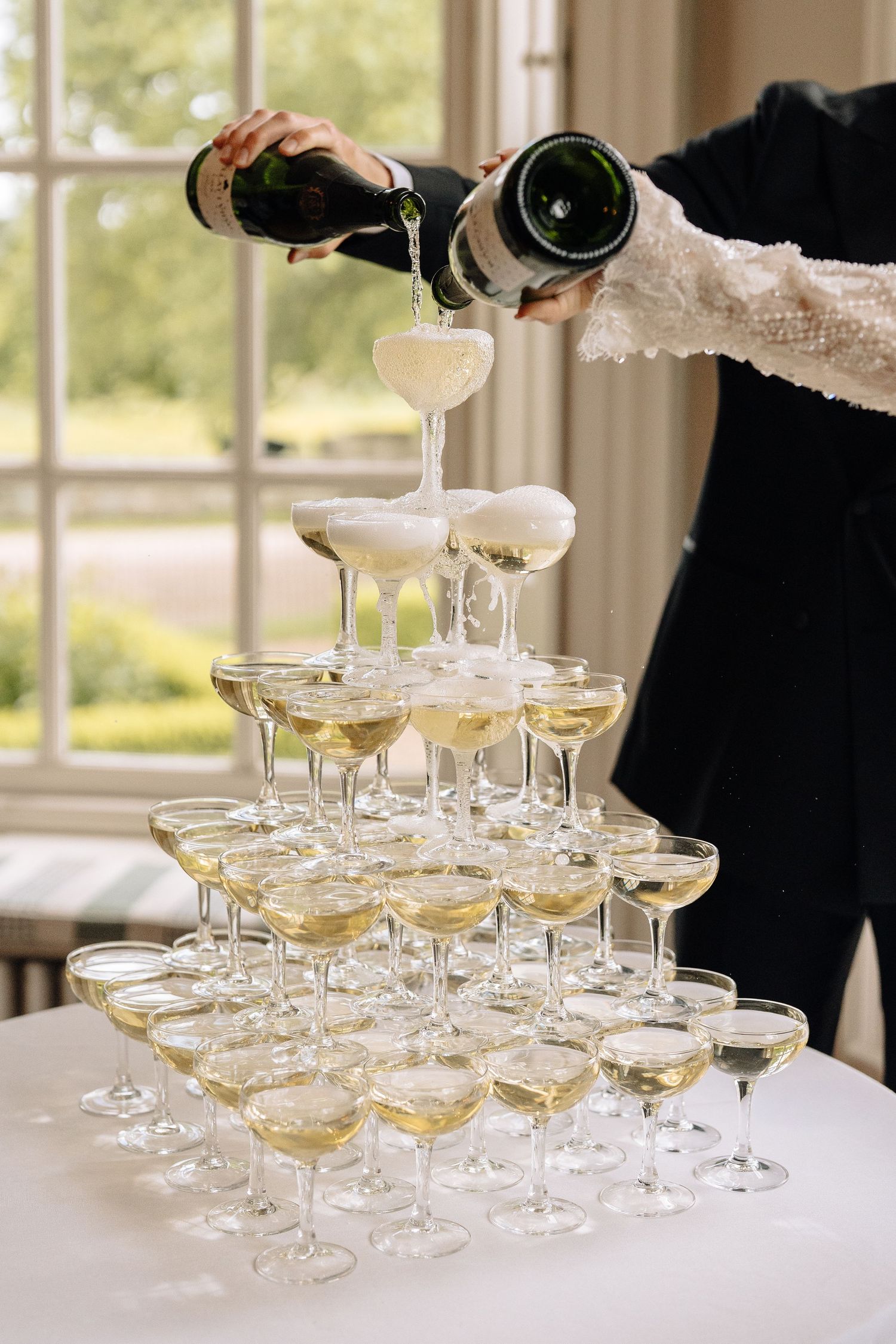 Two people pouring champagne into a elegant pyramid tower of coupe glasses at a wedding reception.