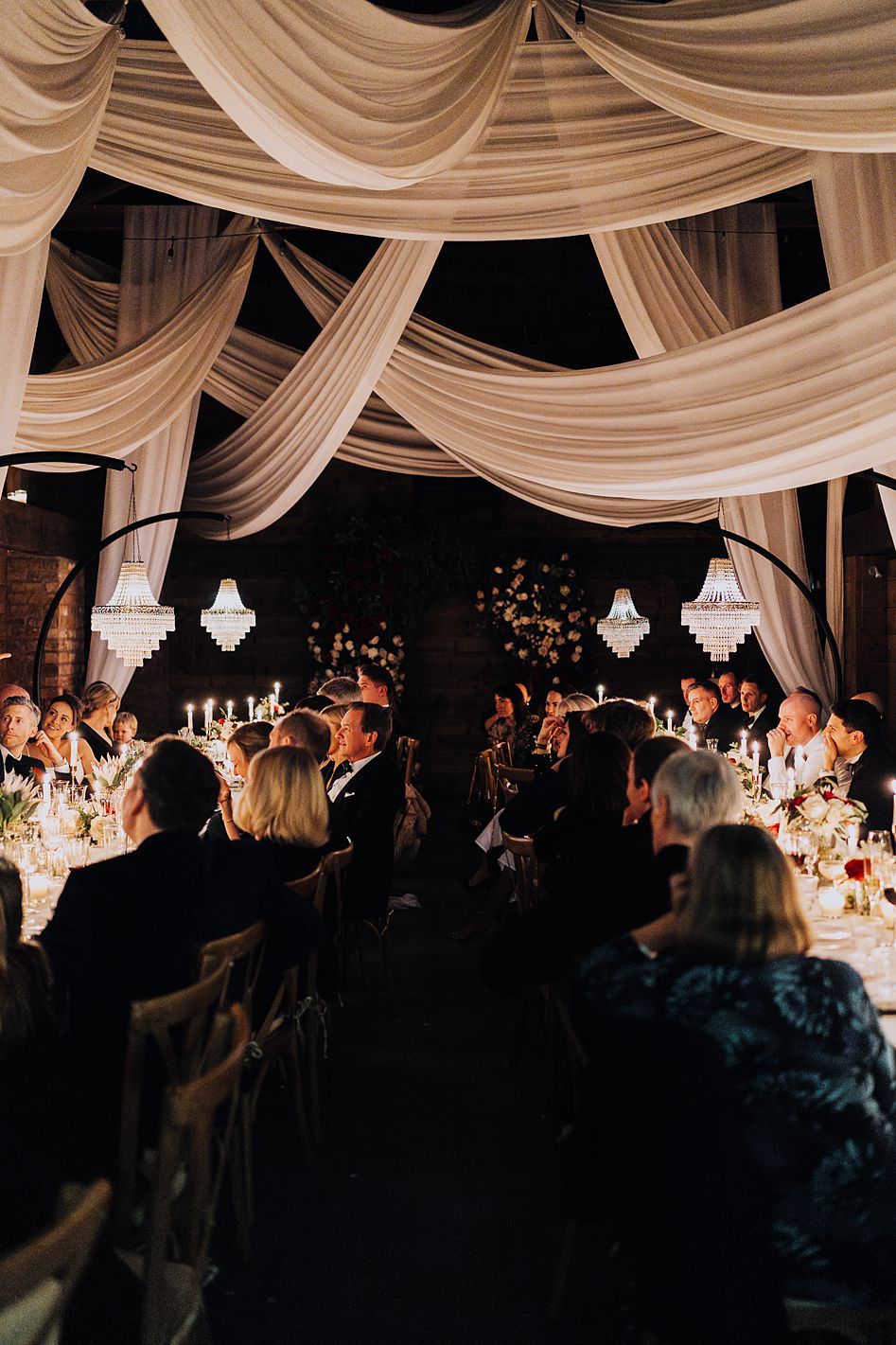 Elegant wedding reception hall with draped ivory fabric ceiling, chandeliers, and candlelit guest tables.