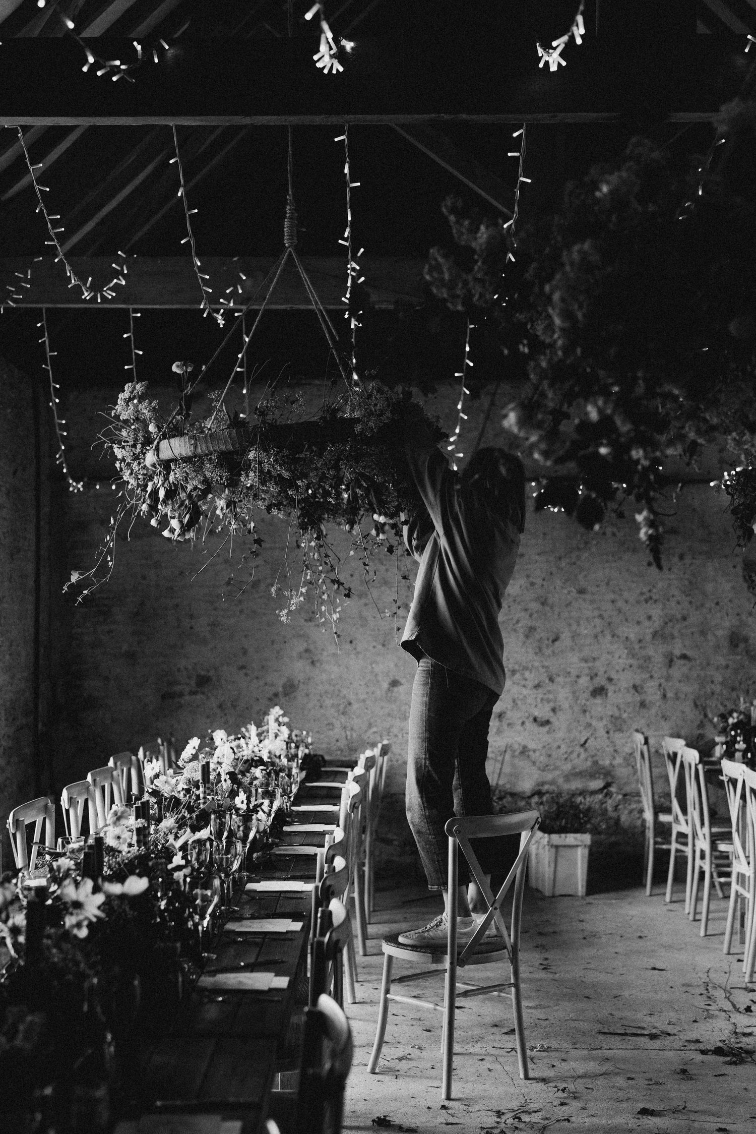 Black and white photo of a person arranging hanging floral installations in a rustic barn venue.