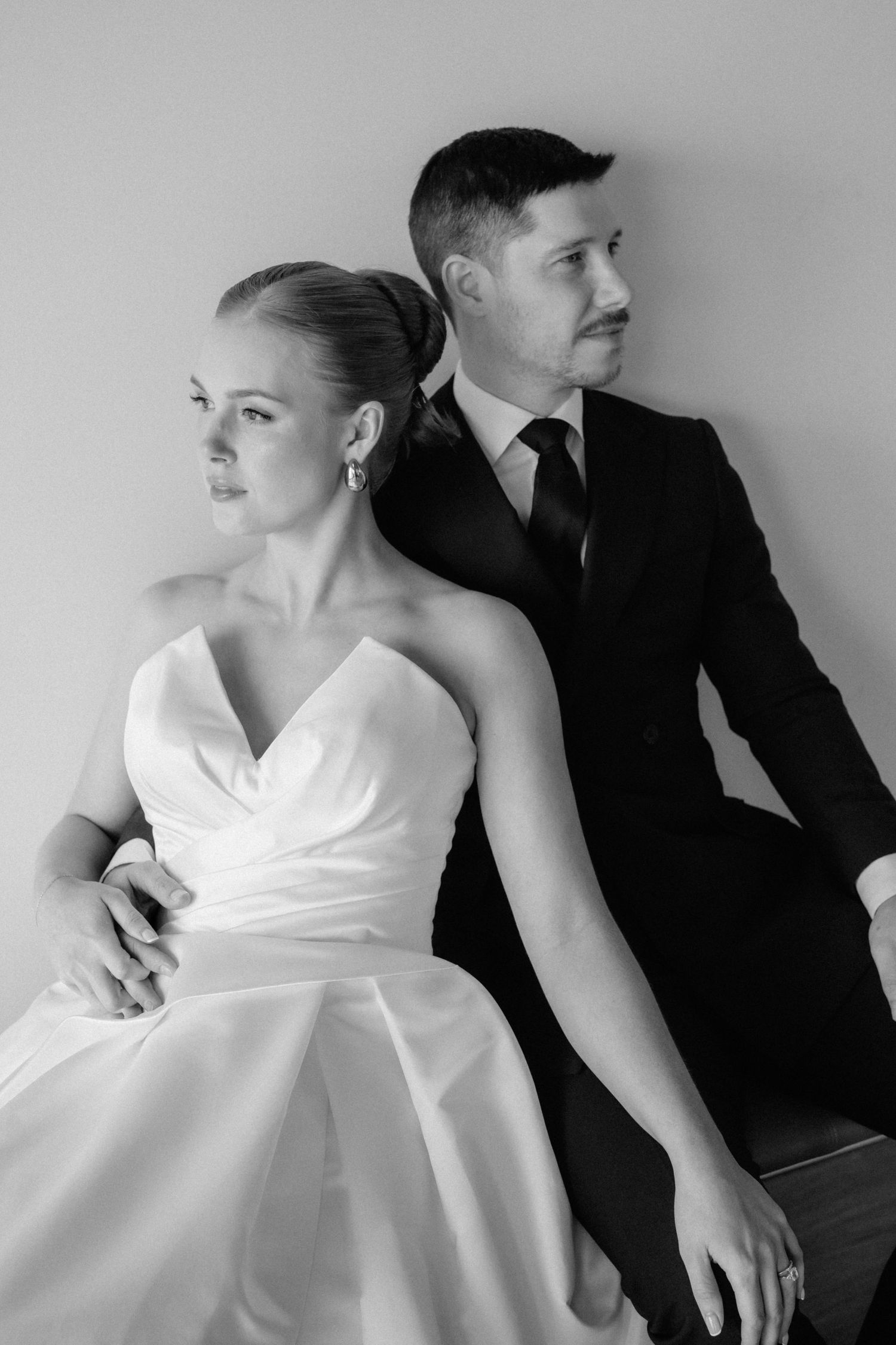 Black and white wedding photo of bride in strapless gown and groom in suit sitting back to back, looking away.