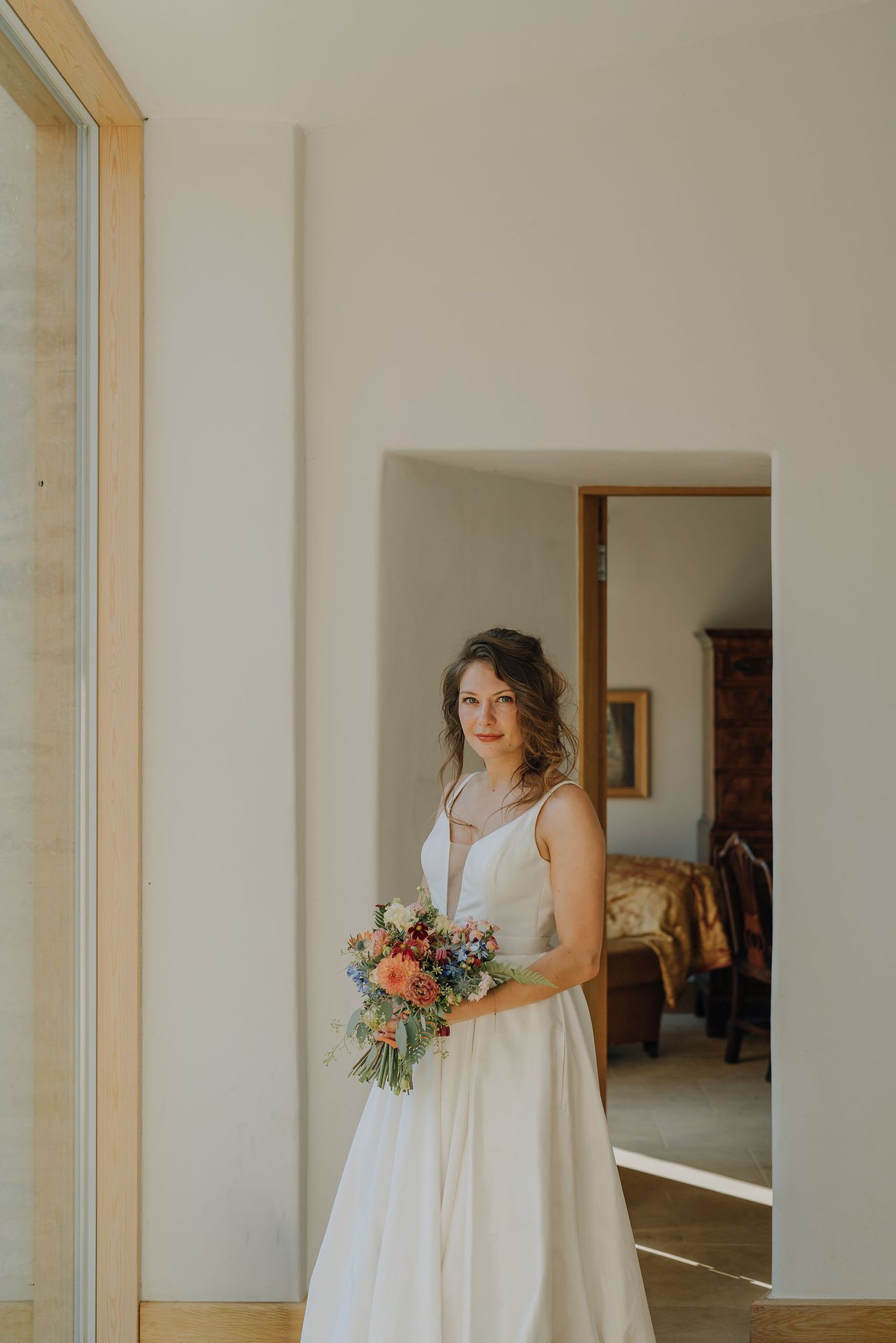 Smiling bride in white gown holding colorful bouquet standing in bright modern doorway.