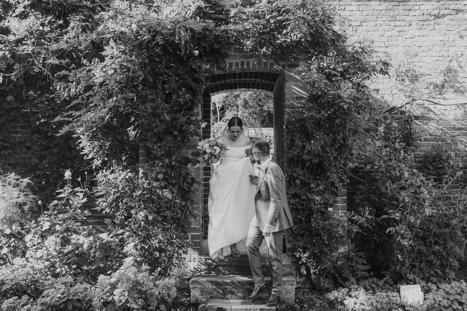 Black and white photo of bride and child standing in ivy-covered stone doorway with bouquet.