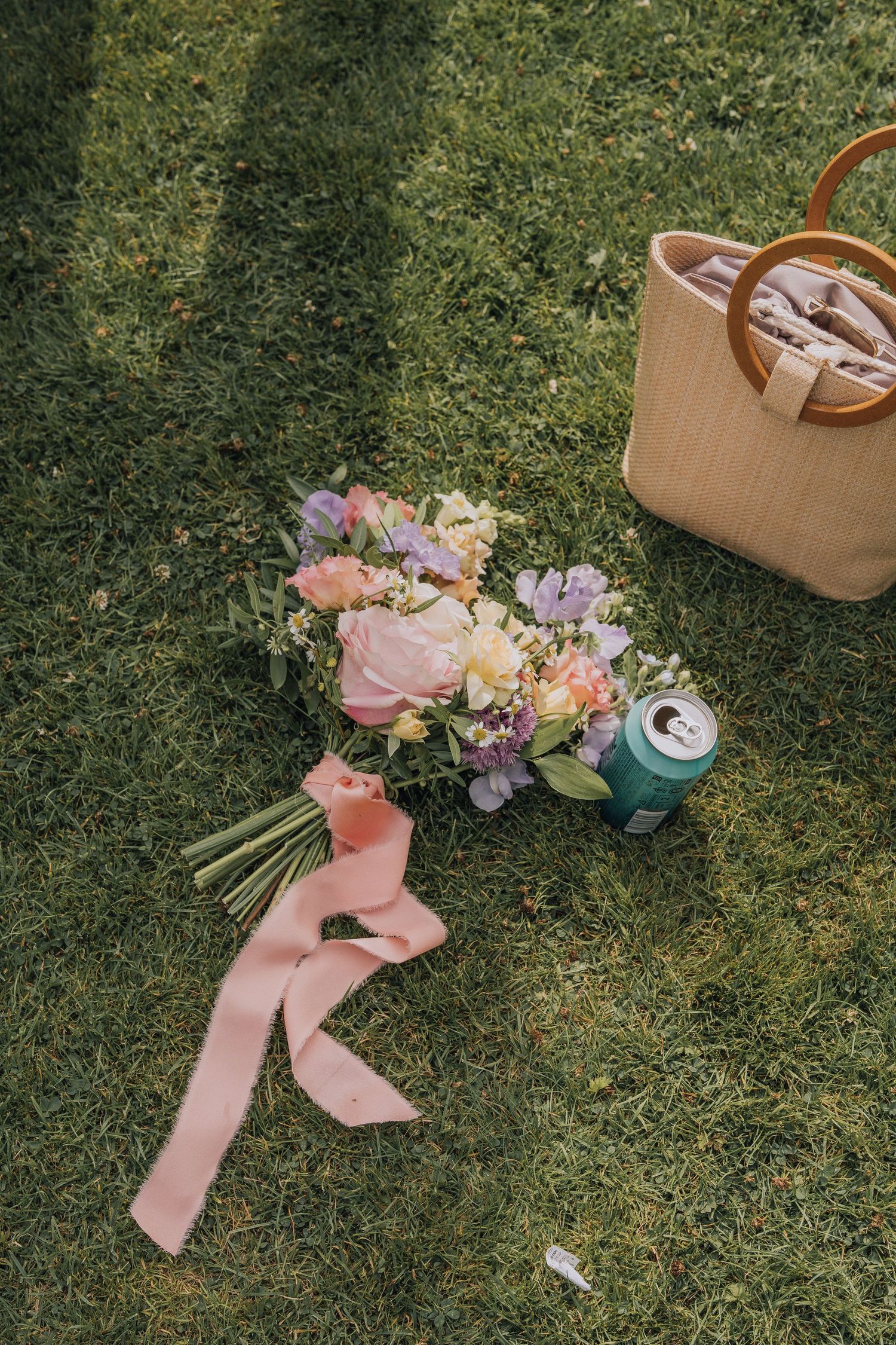Colorful bridal bouquet with pink ribbon lying on grass beside wicker bag and beverage can.