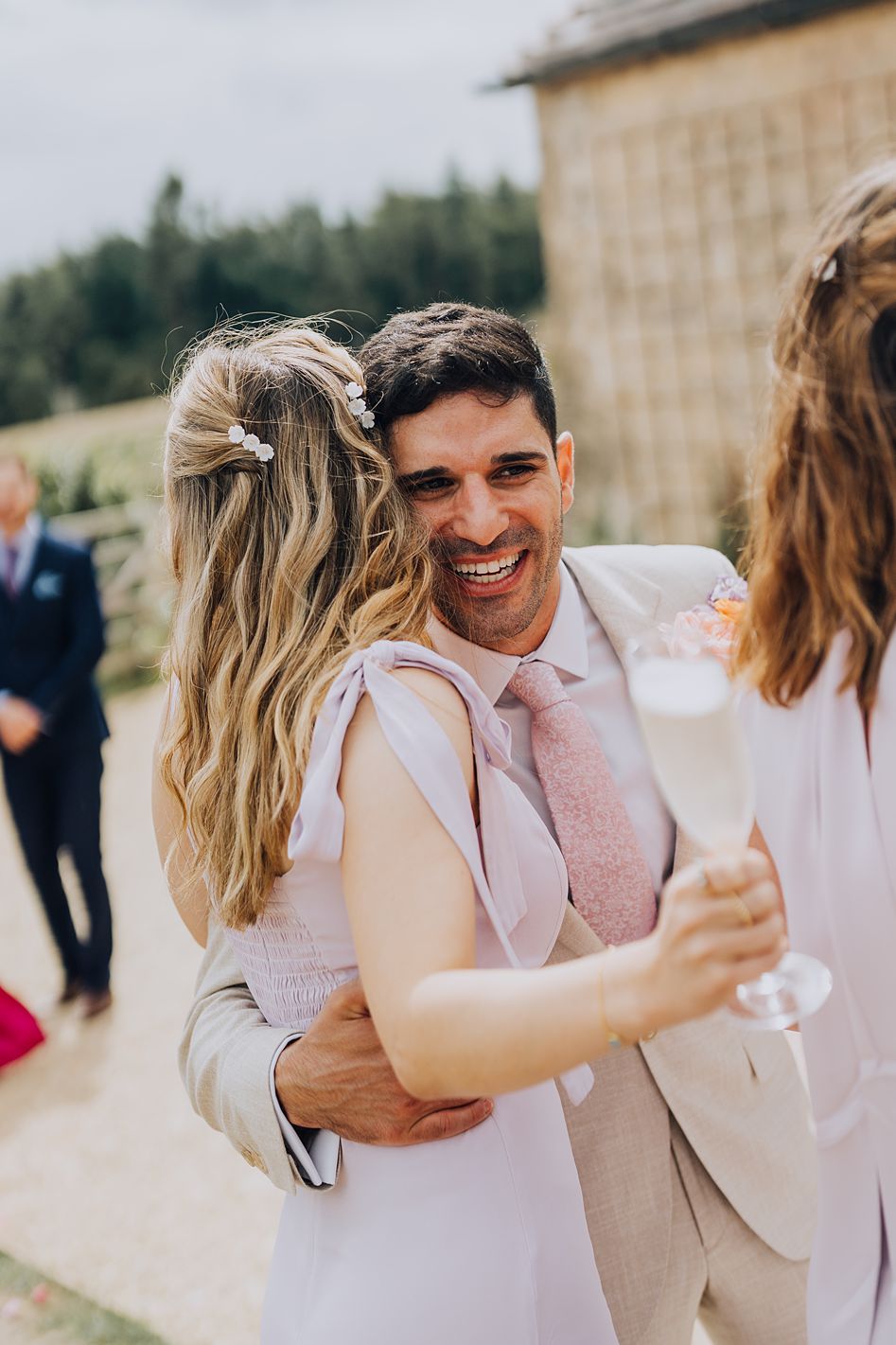 Joyful groom in beige suit hugging guest while holding champagne flute at outdoor wedding reception.