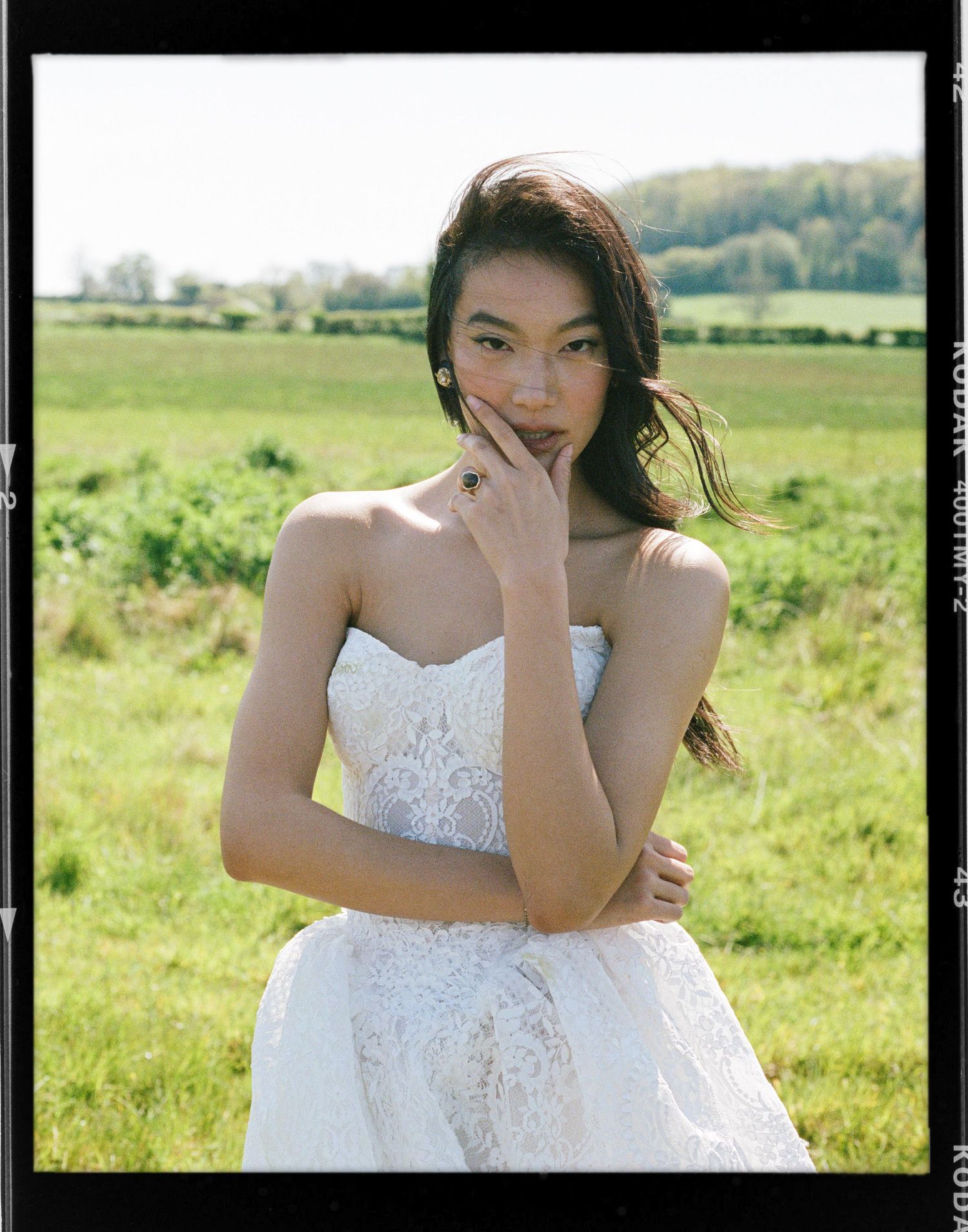 A young woman in a strapless white lace gown poses gracefully in a lush green field on a sunny day. Taken on 35mm film