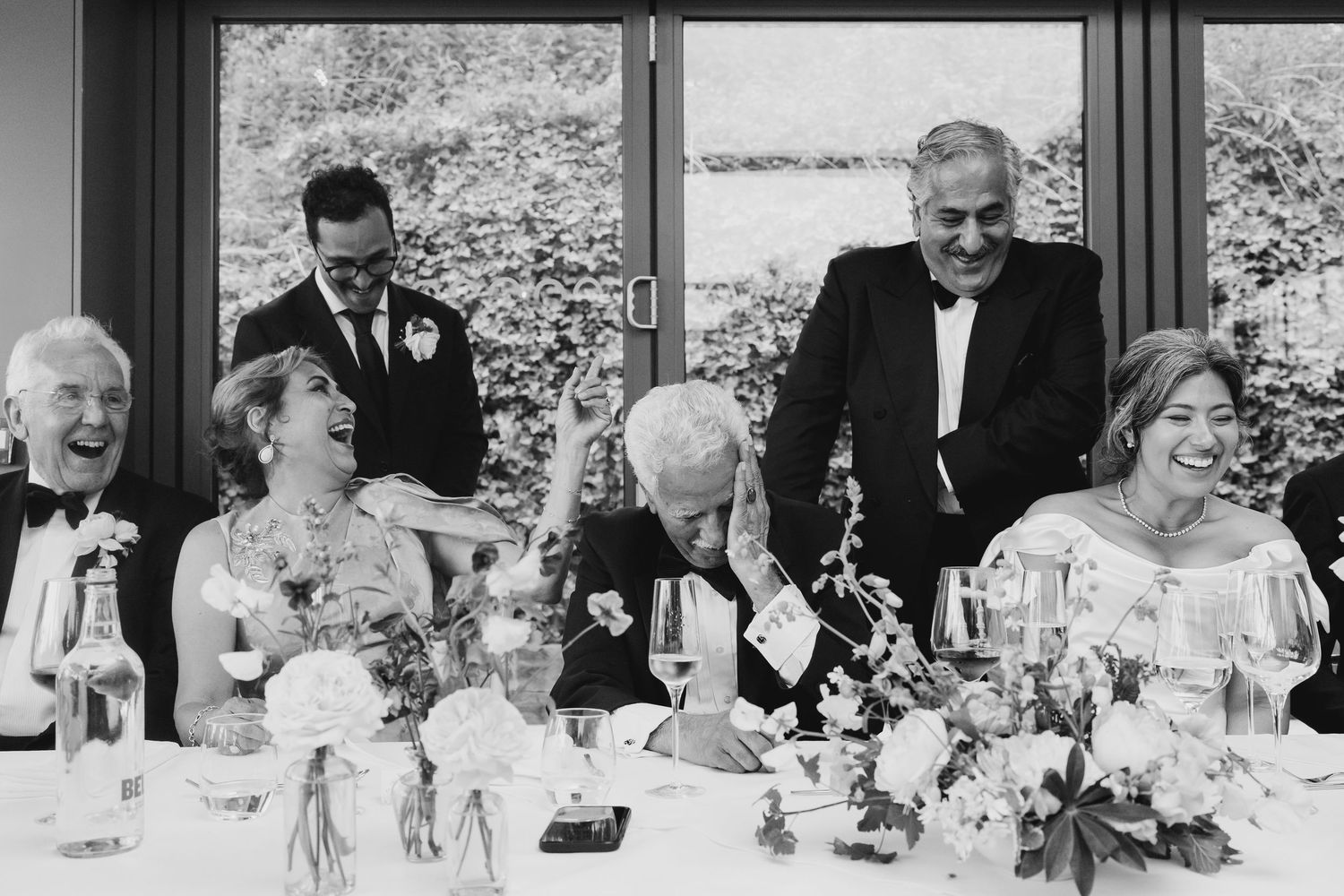 Wedding party laughing joyfully at the head table during reception speeches in a bright venue.