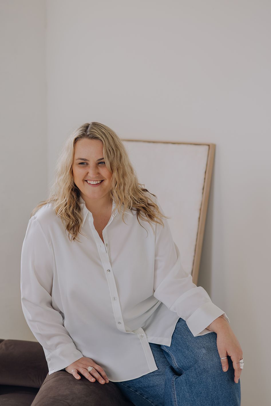 Smiling blonde woman in white shirt sitting casually against a white wall in a bright room.