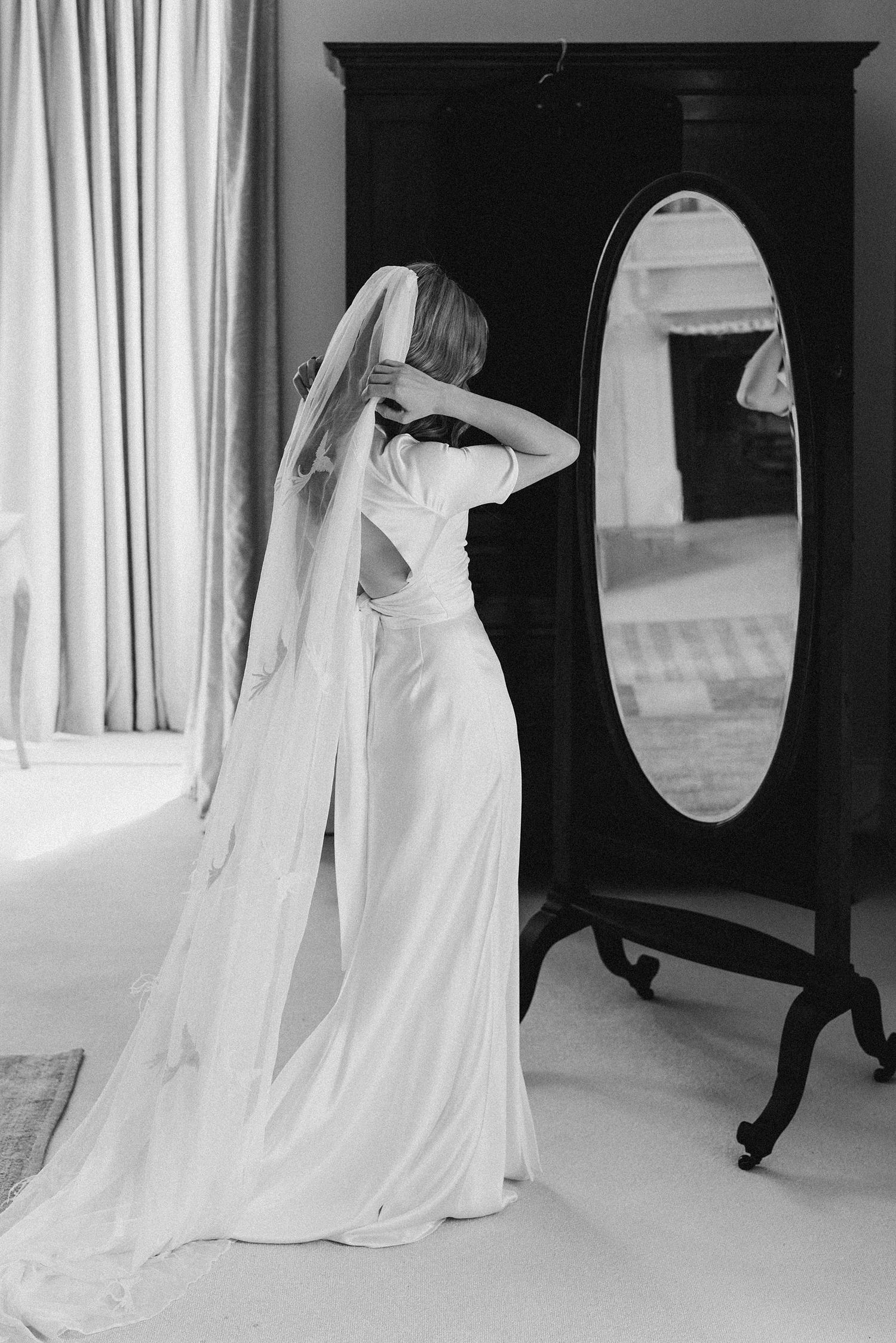Bride in elegant white gown and veil adjusting hair before antique mirror in black and white photo.