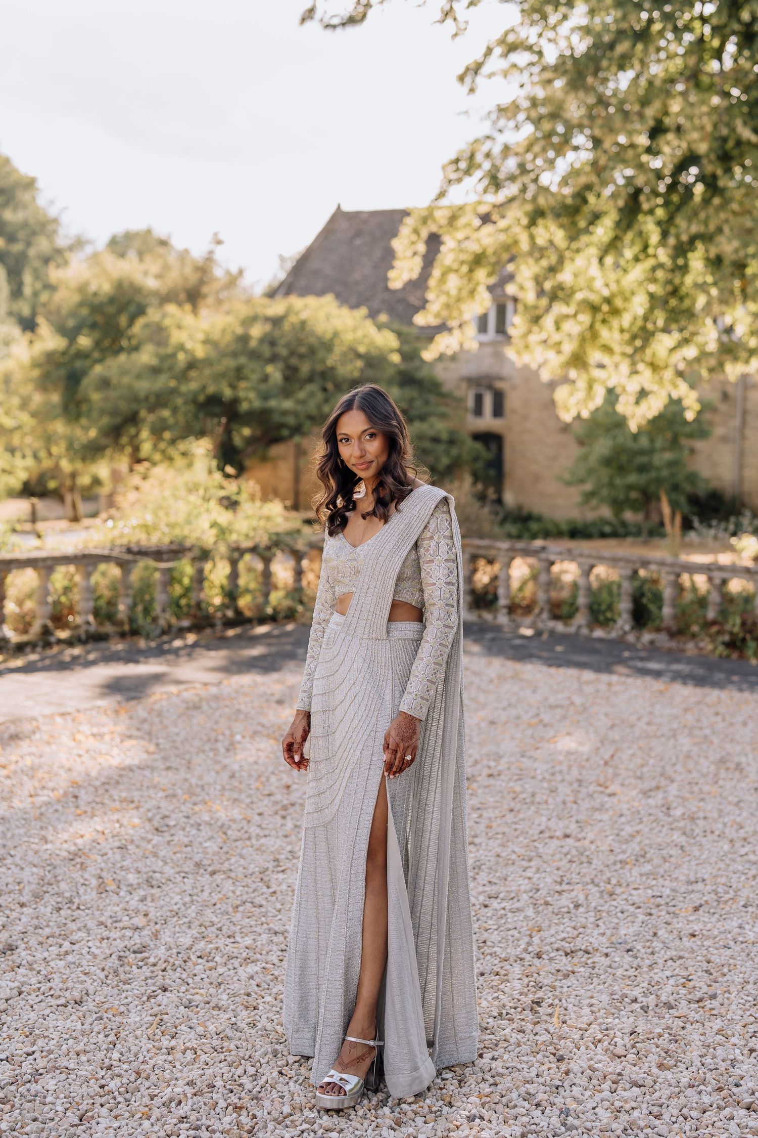 Bride poses in silver embellished saree gown with cape on gravel path outside stone manor house.