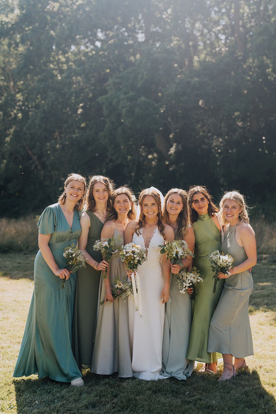Bride with six bridesmaids in mismatched sage and green dresses holding wildflower bouquets outdoors.
