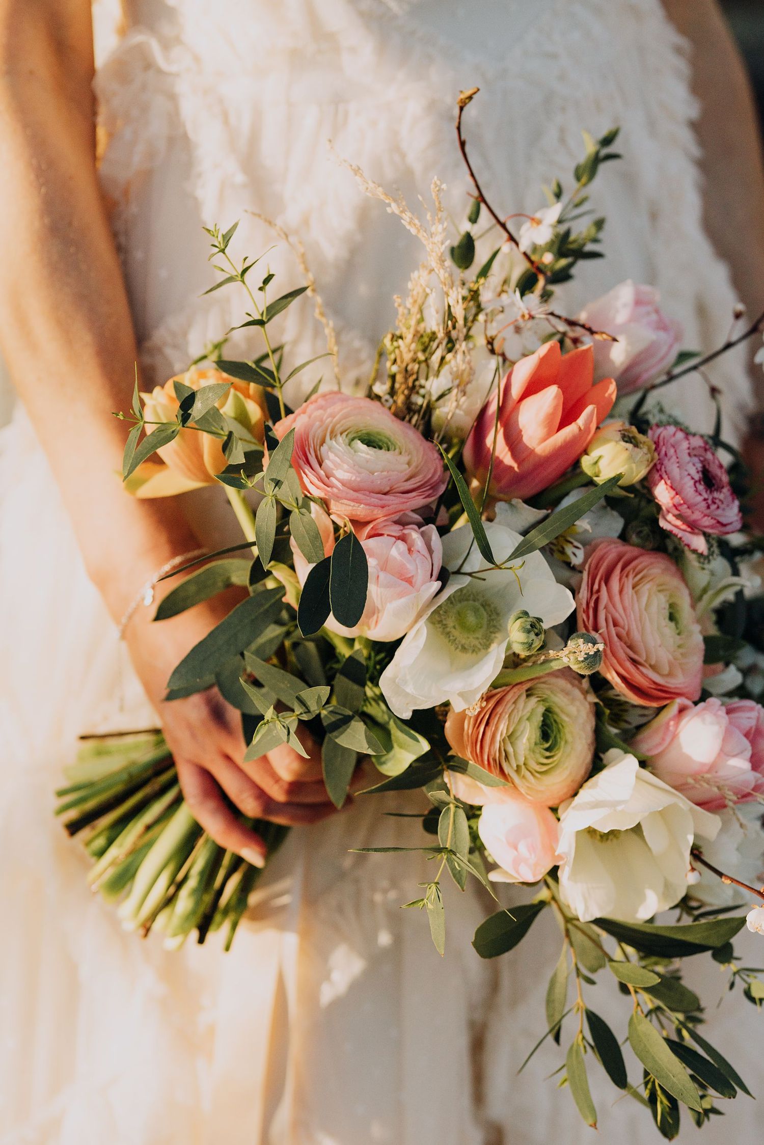 Bride holds a lush spring wedding bouquet of pink ranunculus, tulips, and white anemones.