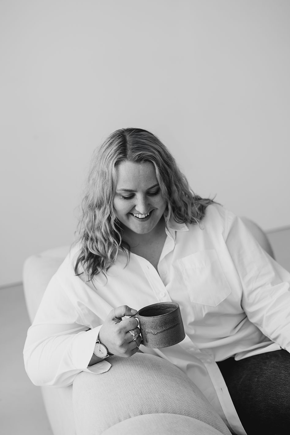 Blonde woman in white shirt smiling while holding a mug on a sofa in black and white.