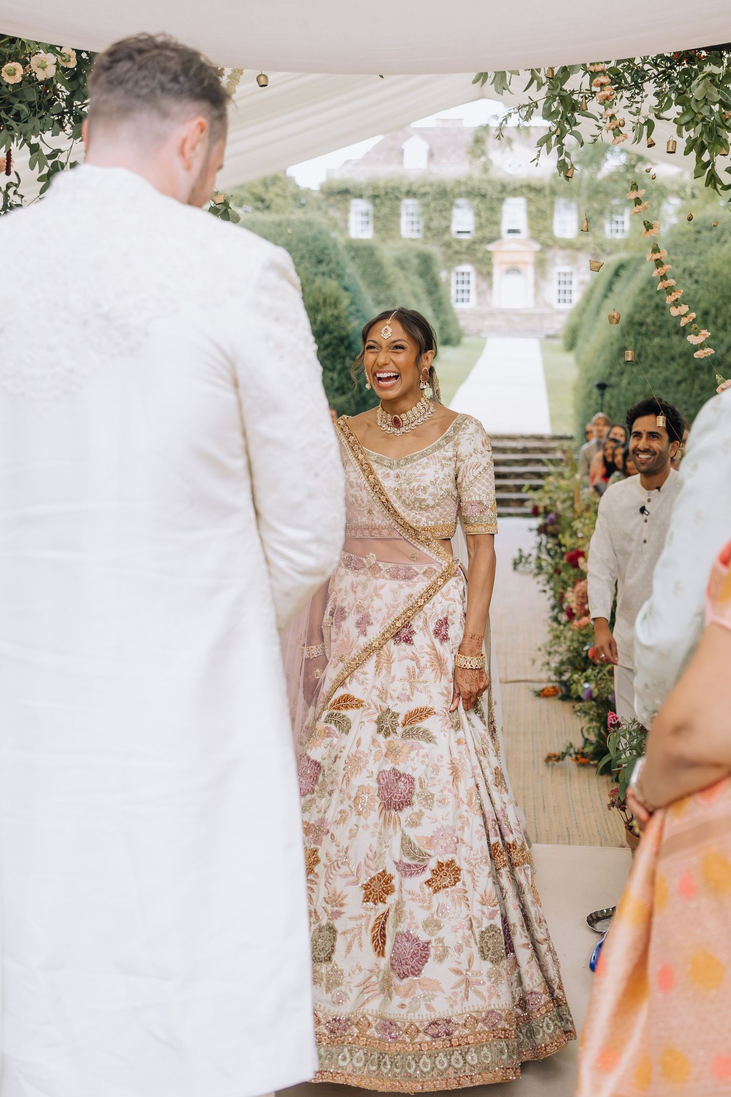 Radiant Indian bride in floral lehenga laughing joyfully as groom approaches during outdoor wedding ceremony.