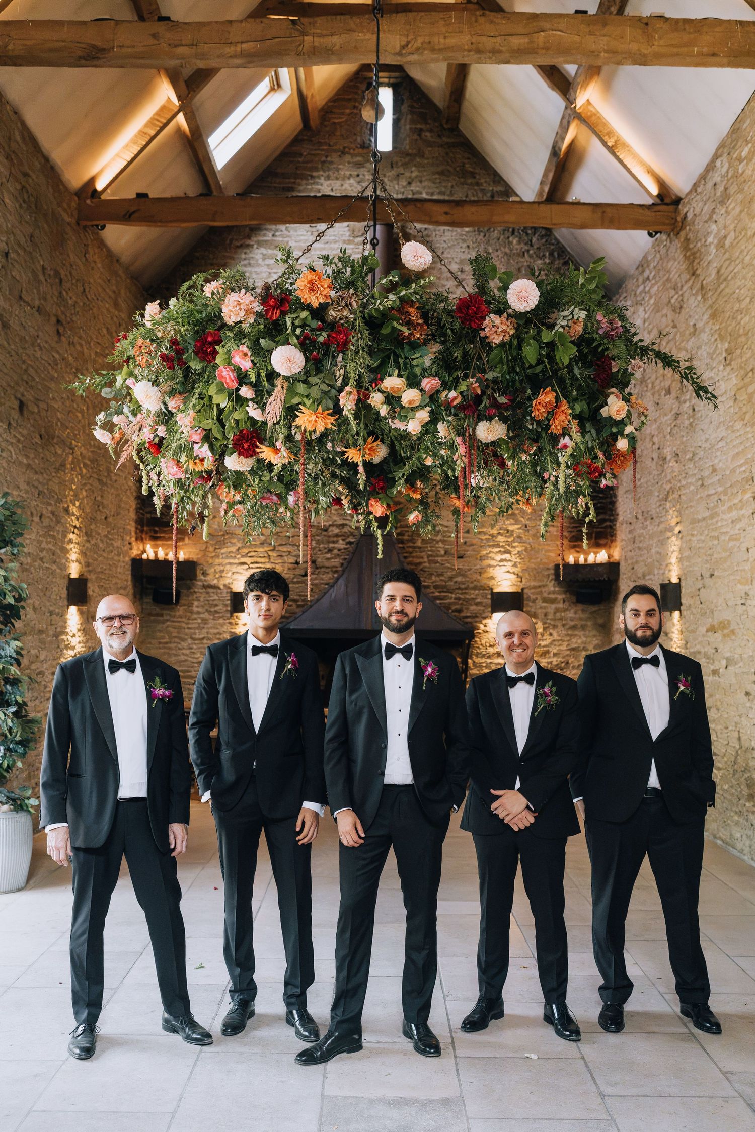 Five groomsmen in black tuxedos pose under a lush floral installation in a rustic barn wedding venue.