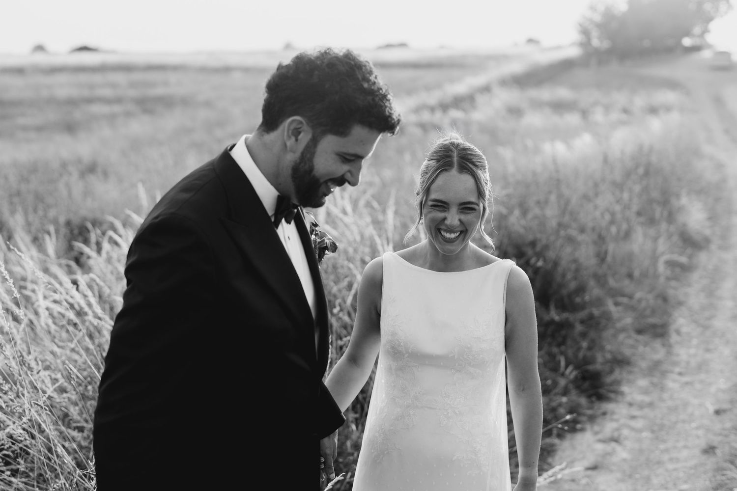 Black and white photo of a smiling bride in a sleek gown and groom in a tuxedo walking together in a field.
